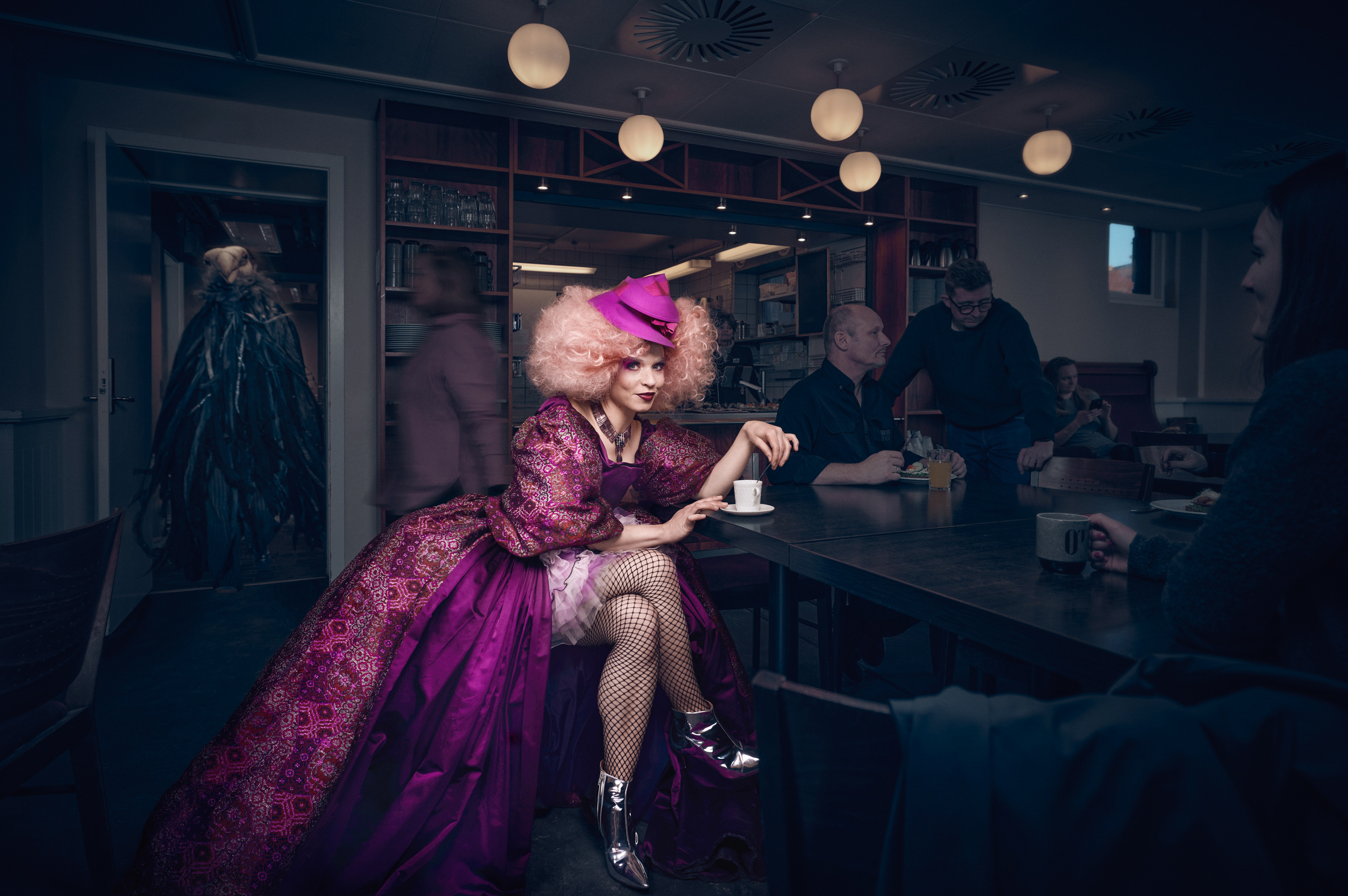 En kvinde klædt i pompøs lilla kjole, stor paryk og pink hat er i front på billedet. Personer i mørke i baggrunden. Odense Teater sæson 26/27