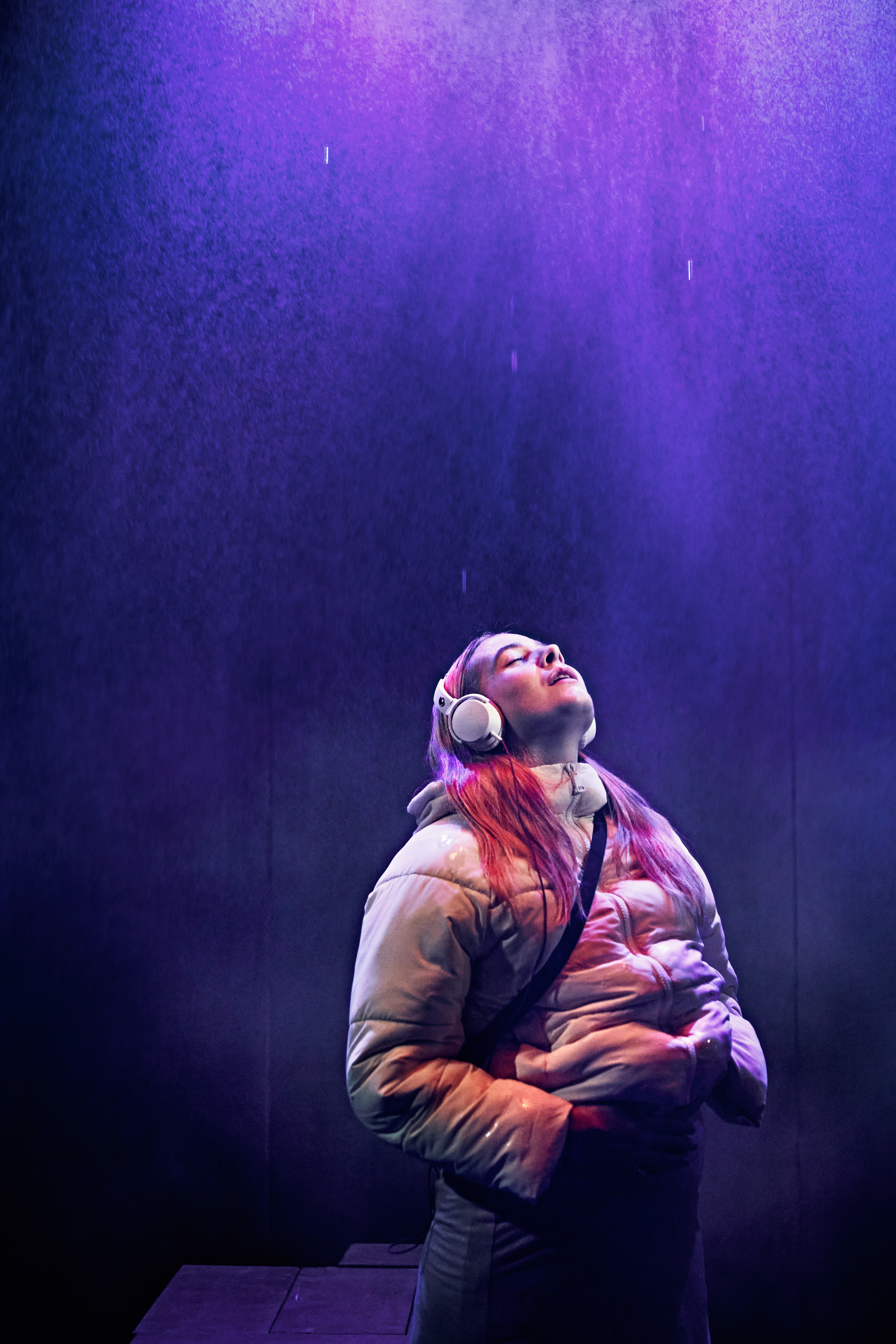 Ung kvinde med langt hår og høretelefoner står i regn med ansigtet vendt op mod himlen. Fra forestilling Maria på Odense Teater