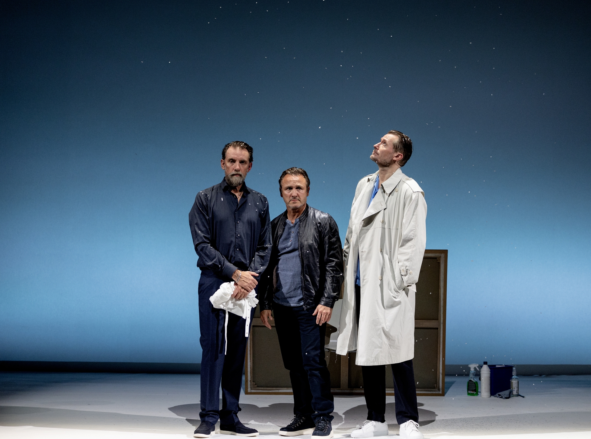 3 mænd i forskellig højde står og ser opgivende ud. Et stort maleri står med bagsiden ud bag dem. Blå baggrund. Fra forestillingen Kunst