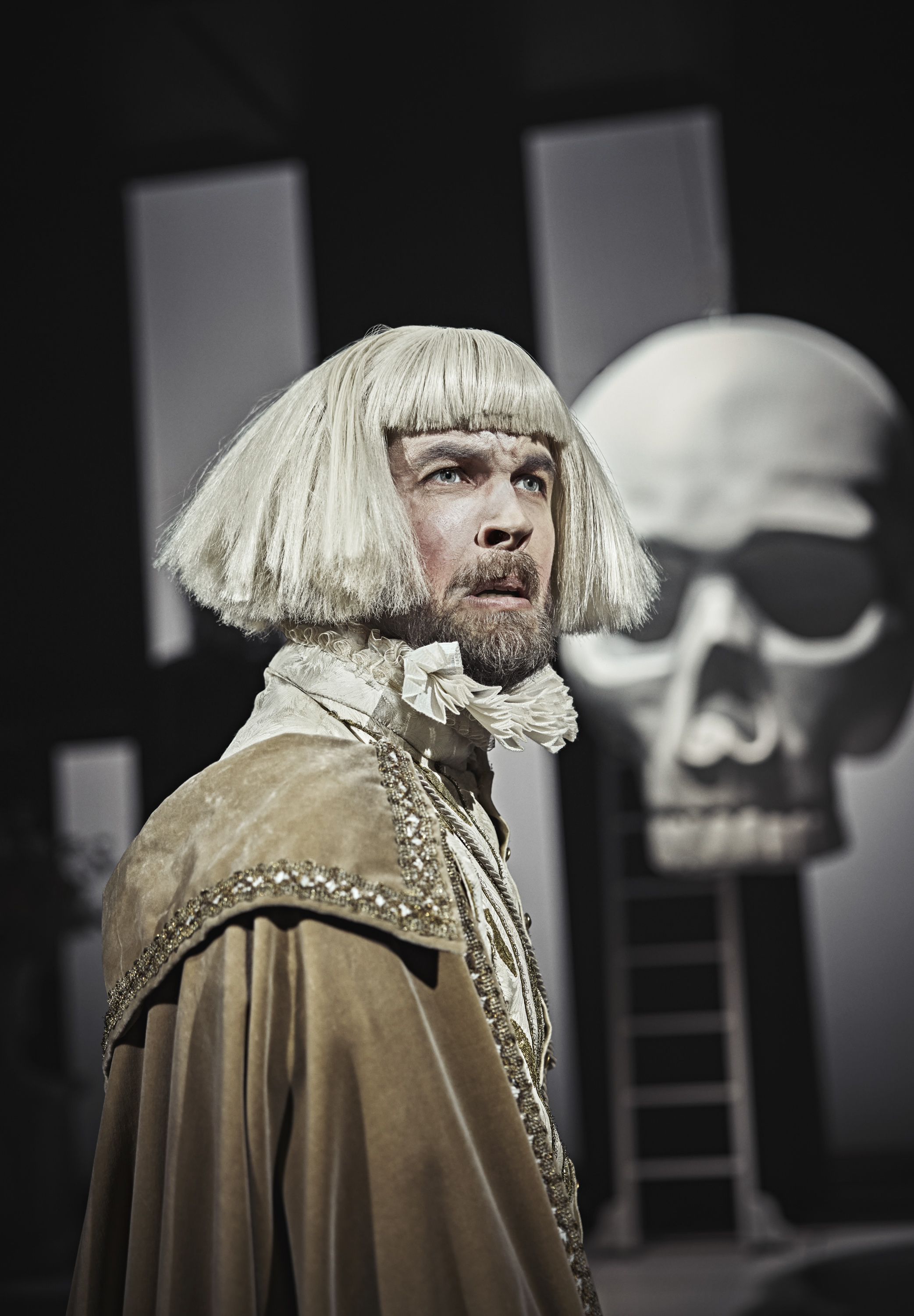 Mand med hvid page paryk og fuldskæg iførtt pibekrave og stor brug velourkappe. Kæmpe hvidt kranie i baggrund. Fra forestilling Cyrano de Bergerac på Odense Teater