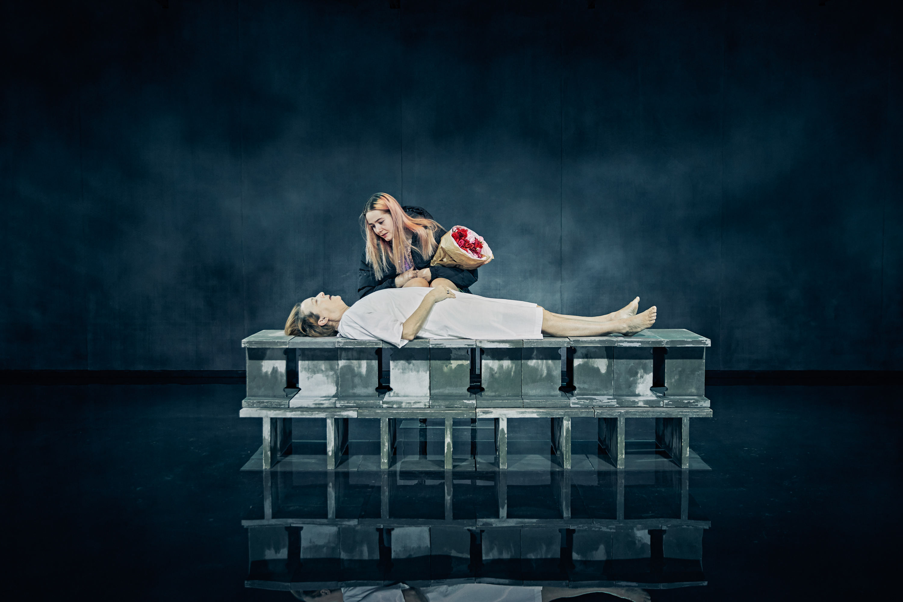 Kvinde ligger på cementblokke iført hvid natkjole. Ung kvinde med en buket blomster står bøjet over hende. Fra forestilling Maria på Odense Teater.