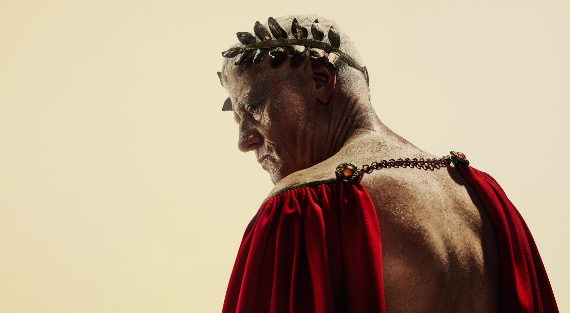 En mand iført rød kappe der blotter hans nøgne ryg og en gylden laurbærkrans på hovedet ser sig over skulderen. Fra forestilling Julius Cæsar på Odense Teater