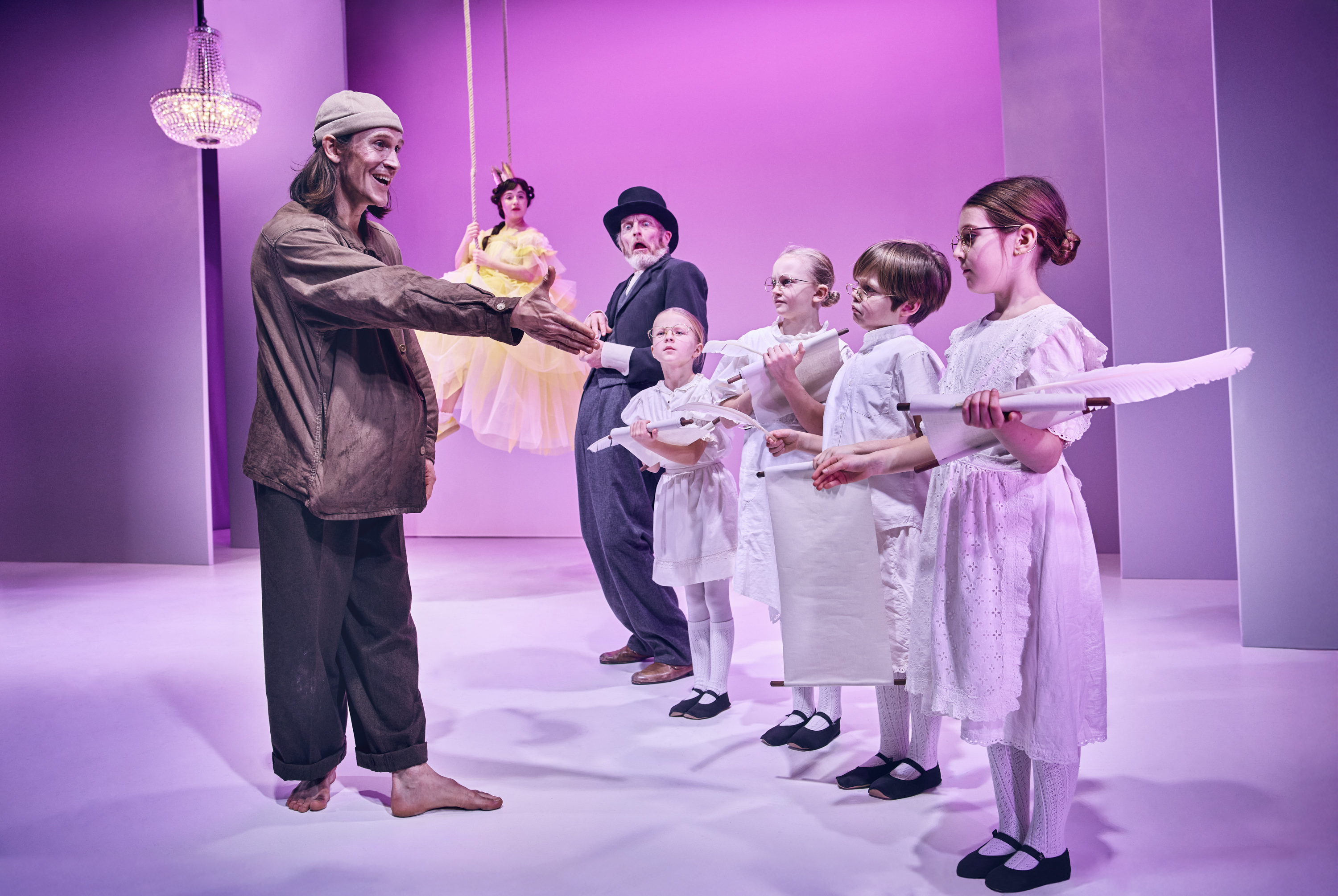 Ung mand med bare fødder hilser på børn klædt i hvidt. I baggrund står ældre mand med hat og prinsesse svæver i luften på gynge. Fra Teaterforestilling Klodshans på Odense Teater