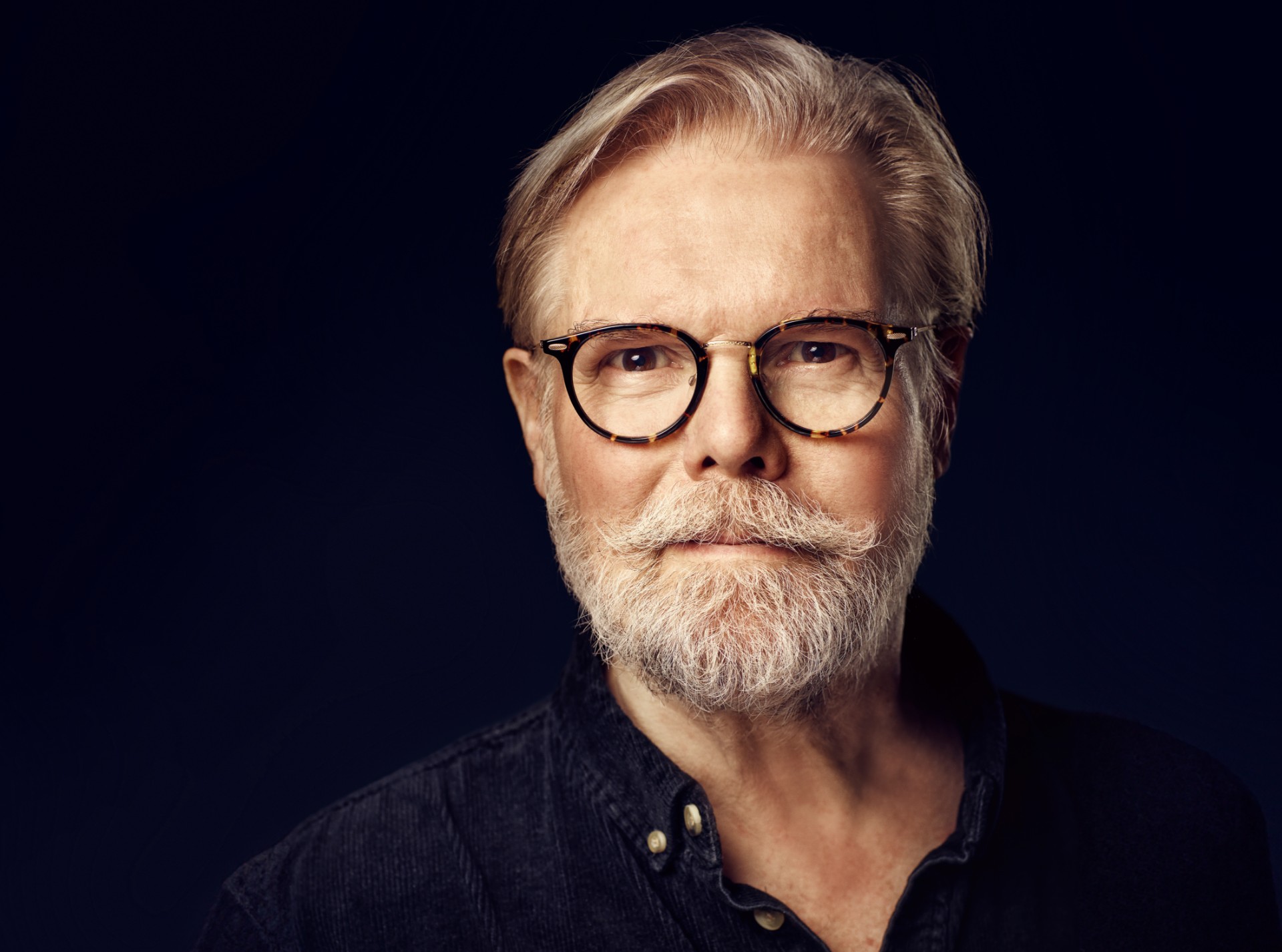 portræt af Anders Gjellerup Koch. Sort baggrund. Mand med gråt hår, fuldskæg, og briller. Iført mørk skjorte.