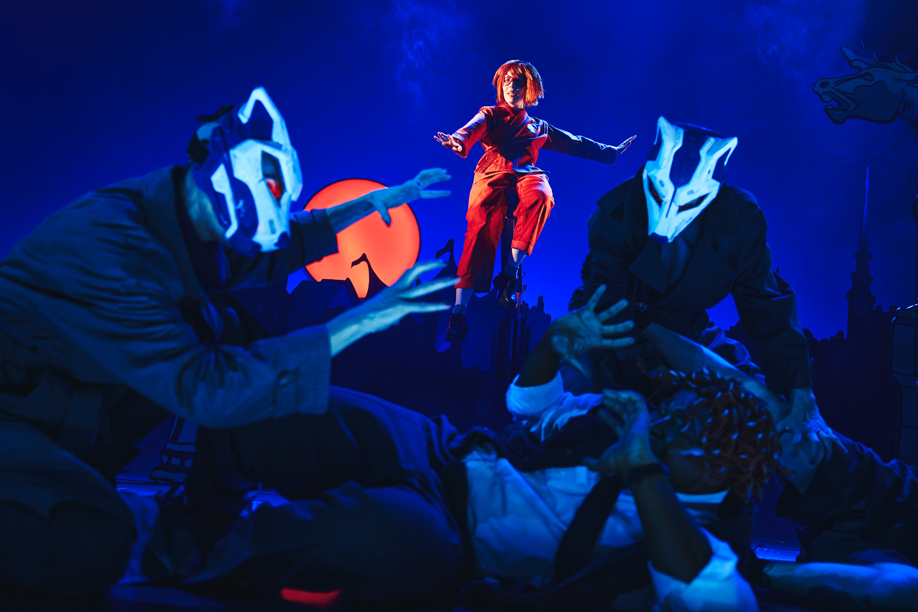 To personer med ulvemasker overfalder en mand der ligger ned. En superhelt i rødt kommer til undsætning. Fra forestillingen Håndbog for superhelte på Odense Teater