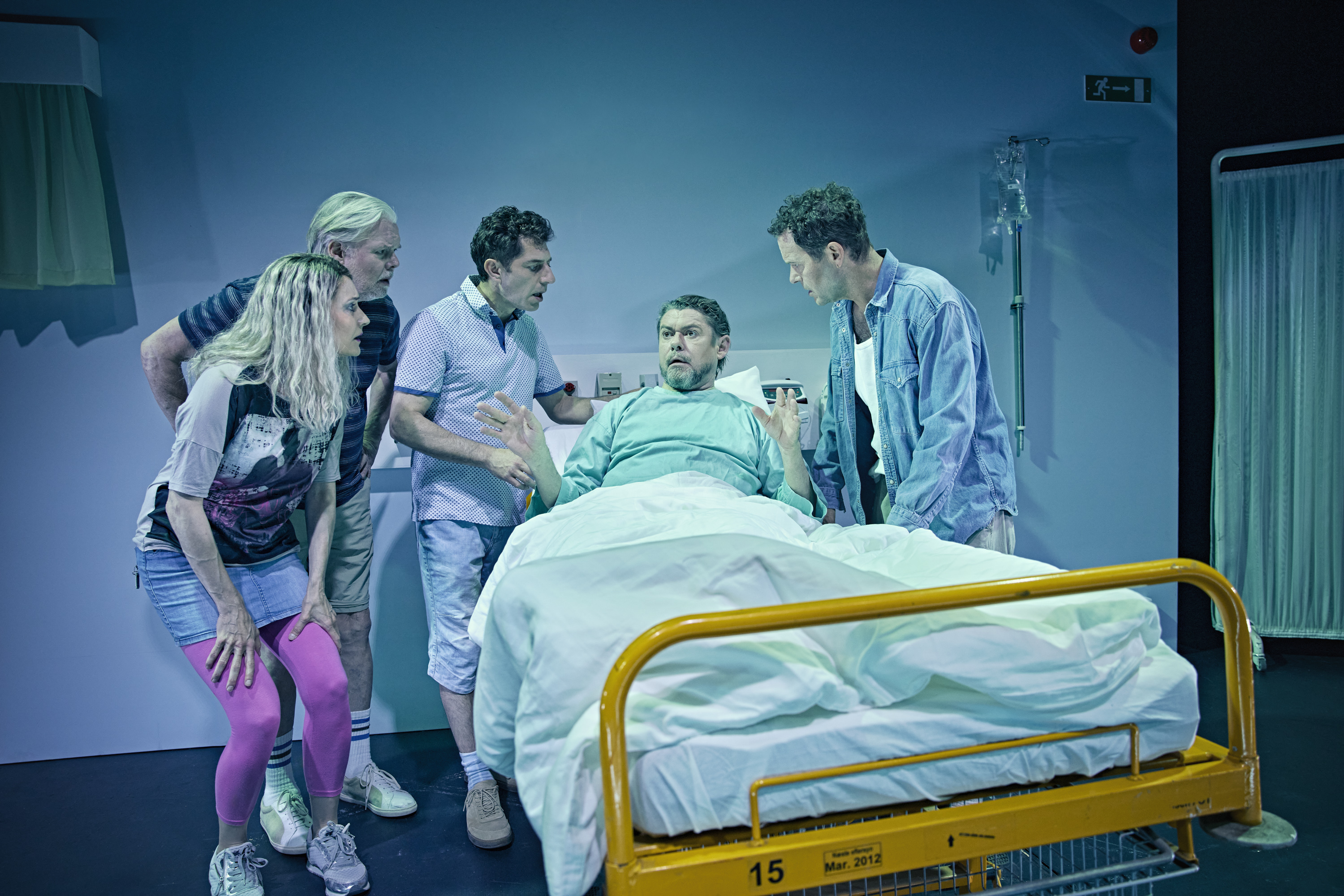 En mand ligger i en gul hospitalsseng. Han er ved at fortælle noget til fire mennesker der står rundt om sengen. Fra teaterforestillingen Miraklet i Madrid på Odense Teater