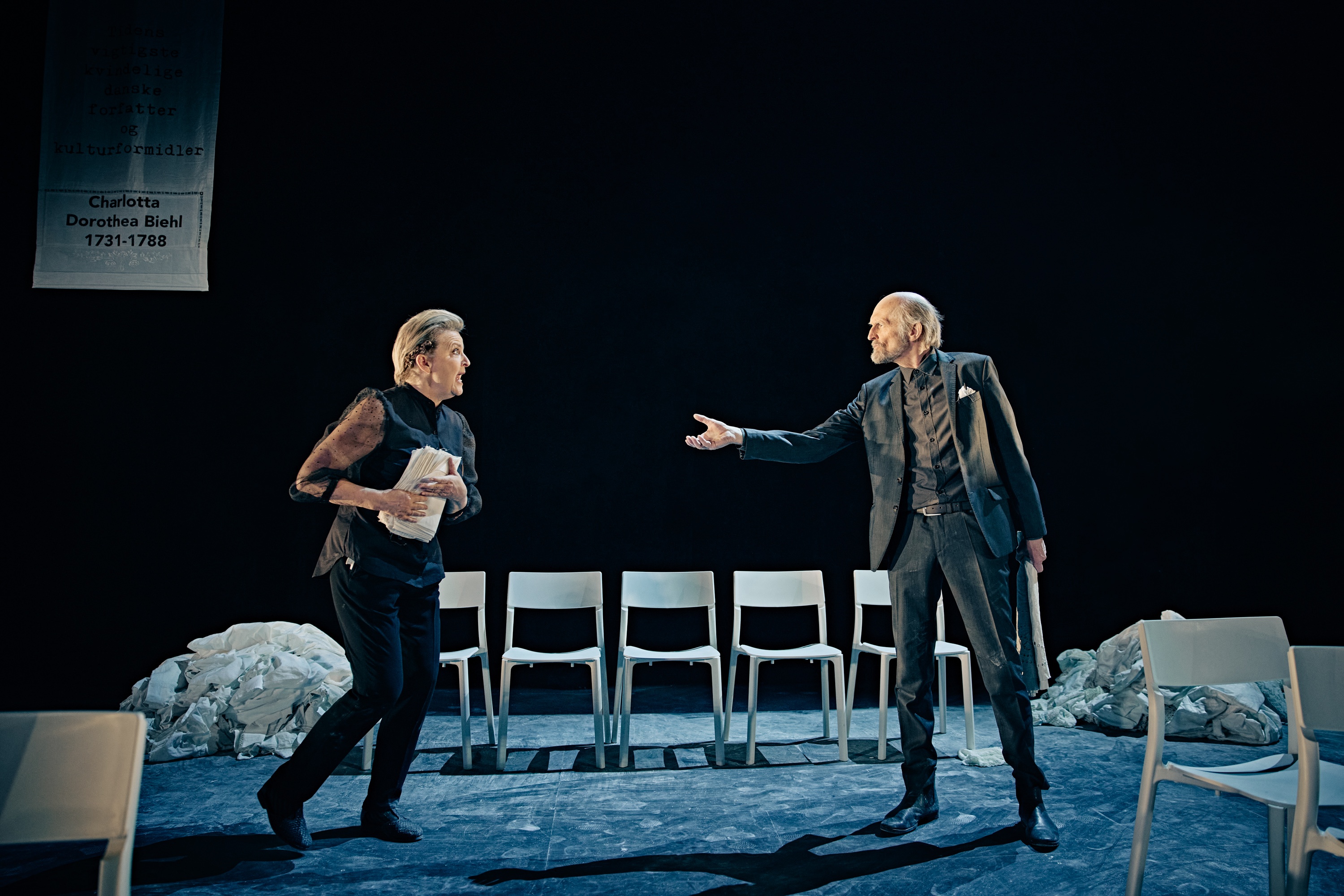 En kvinde og mand på sort scene med hvide stole. Fra forestilling Kvinde kend din historie på Odense Teater