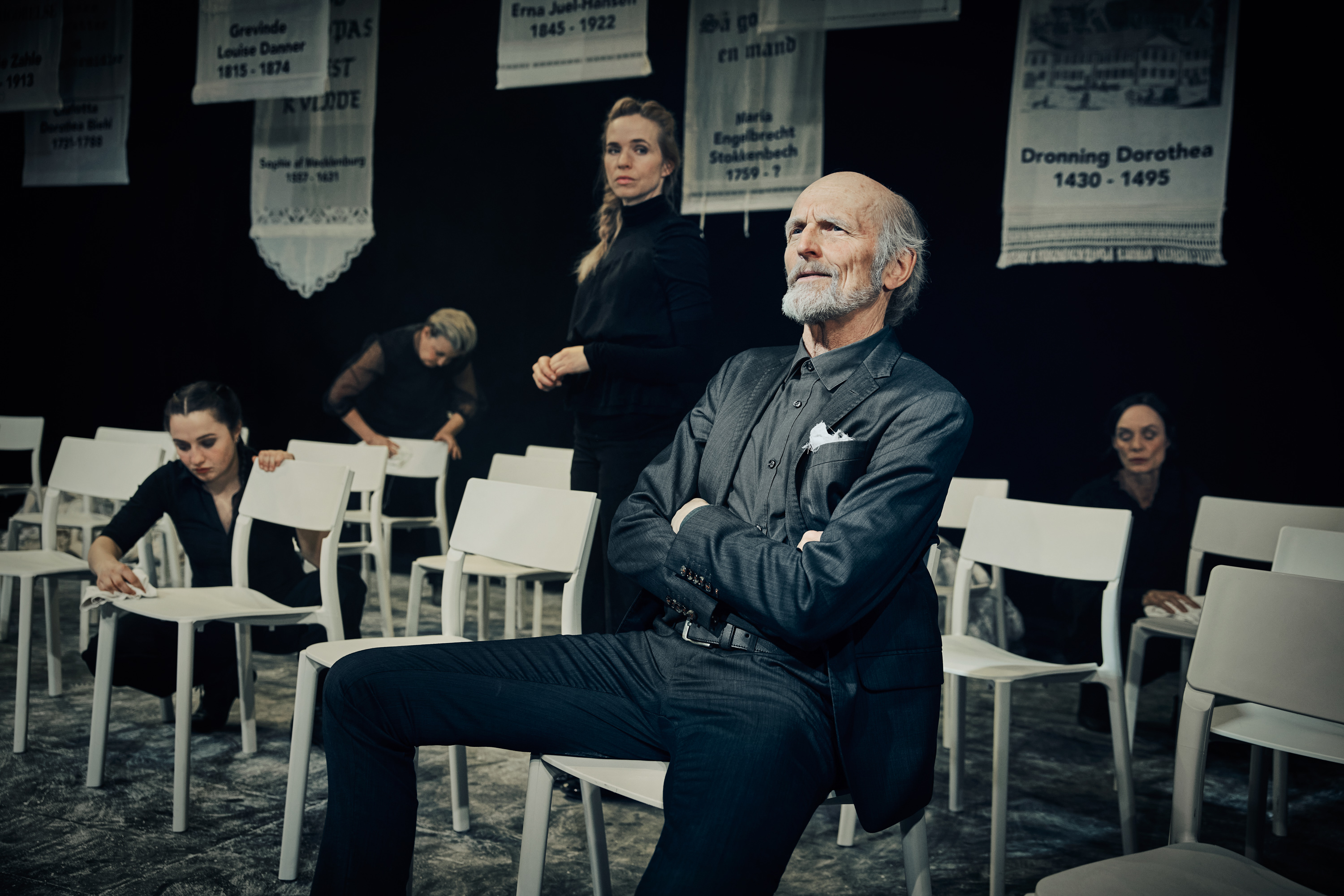 Mand i sort sidder med eftertænksomt udtryk på en hvid stol blandt mange. Fire kvinder er i gang med rengøring. Fra forestilling Kvinde kend din historie på Odense Teater