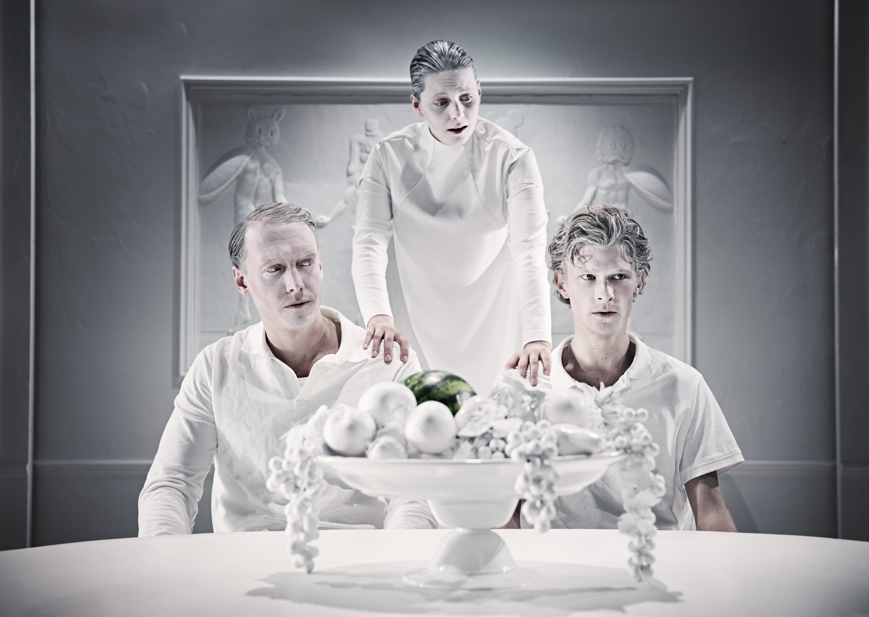 To mænd klædt i hvidt sidder ved bord. En kvinde klædt i hvidt står bagved med en hånd på hver skulder. Fra forestilling Resten kender du fra film på Odense Teater