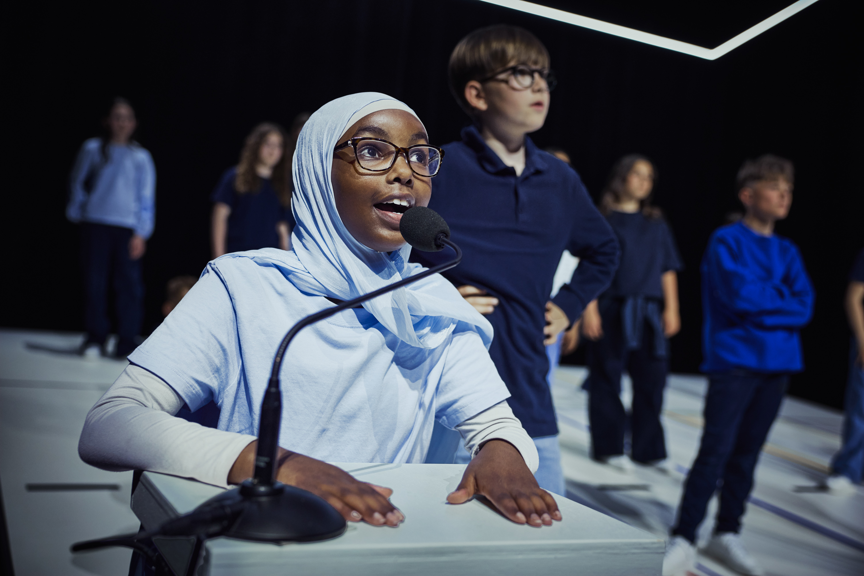 Mørk pige med hijab og briller sidder ved bord og taler i mikrofon. Der er flere børn i baggrunden. Fra forestillingen Mod alle odds på Odense Teater