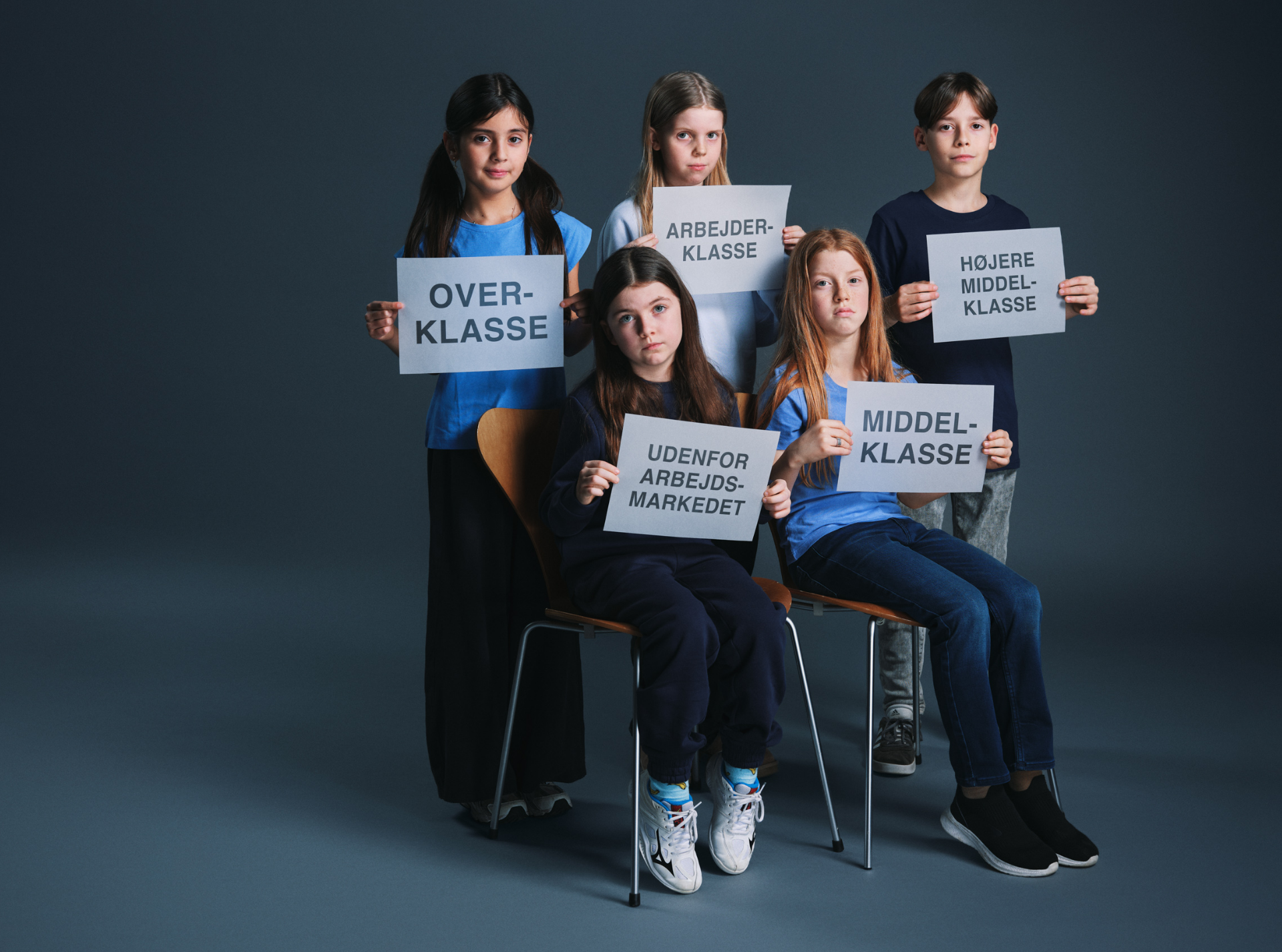 fem børn klædt i blåt. 2 sidder og 3 står. De holder hvide skilte med sort skrift. Fra forestilling Mod alle ods på Odense Teater
