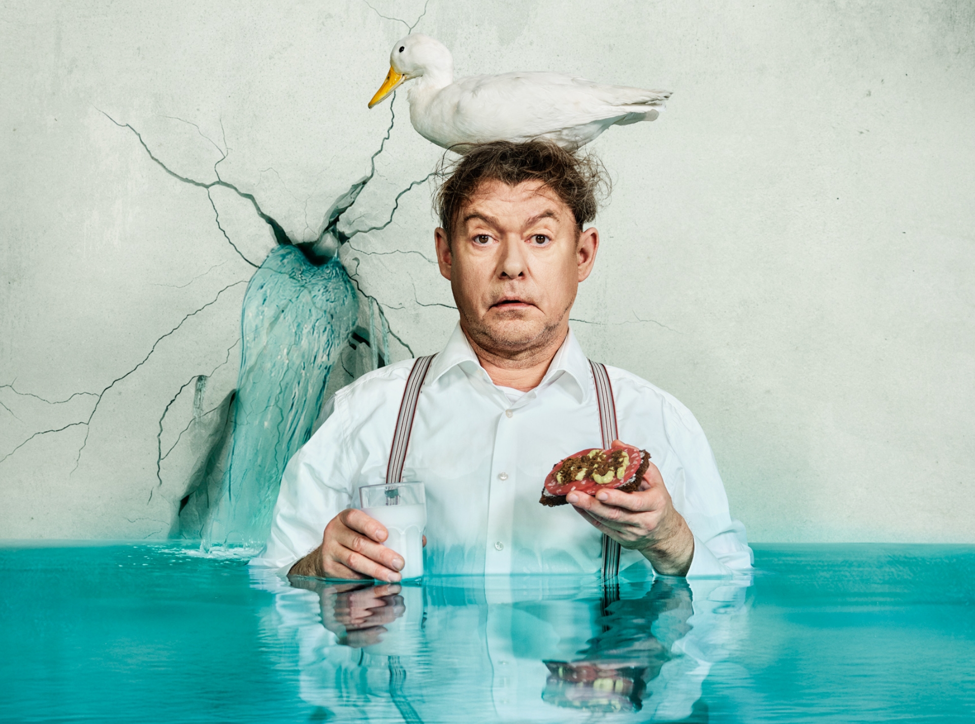 En mand står i vand til livet. Han har en hvid and på hovedet og en spegepølsemad og et glas mælk i hænderne. Fra forestillingen Vi bygger et dige på Odense Teater