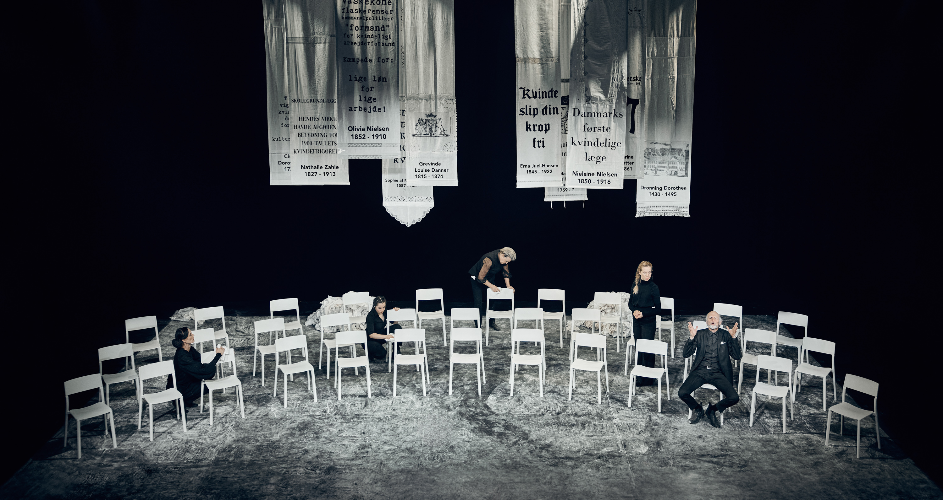 Fire kvinder og en mand og mange hvide stole. Hvide bannere hænger fra loftet med tekst om historiske kvinder. Fra forestilling Kvinde kend din historie på Odense Teater
