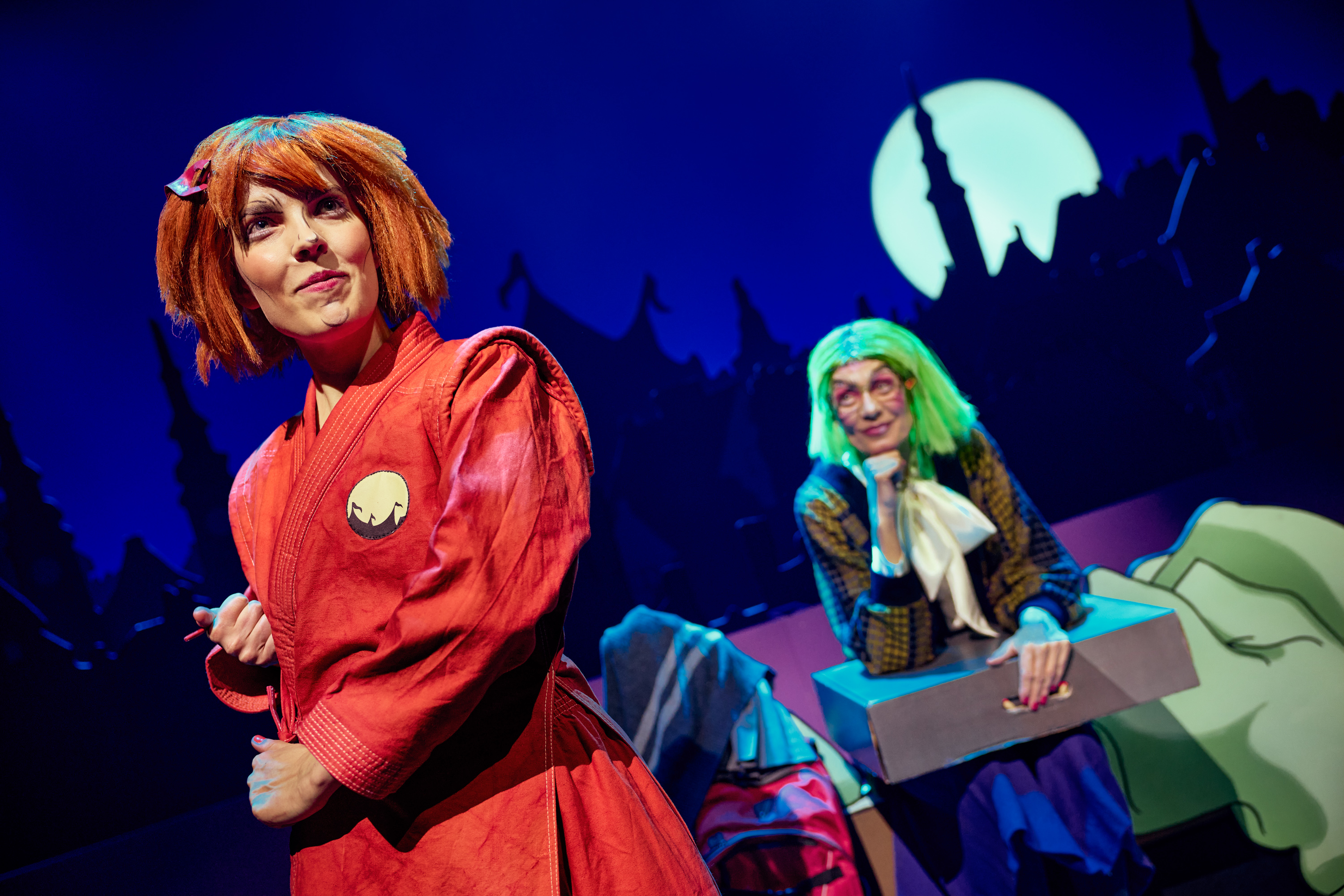 En pige med orange hår i rødt kamptøj. I baggrund en kvinde med grønt hår holder en kasse. Fra forestillingen Håndbog for superhelte på Odense Teater