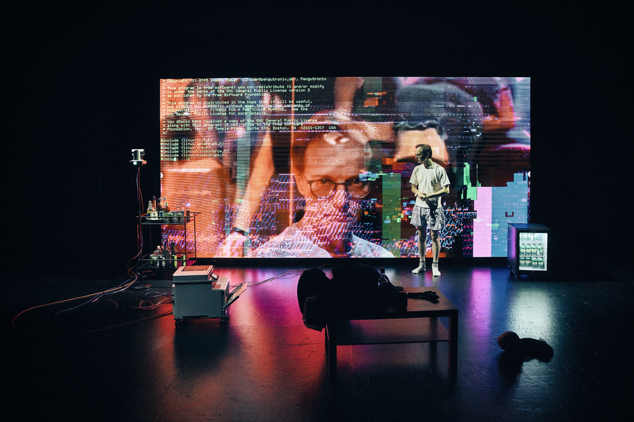 En mand i shorts og t-shirts på en scene. Står foran en stor skærm med nærbillede af hans ansigt. Fra forestillingen AI know you på Odense Teater