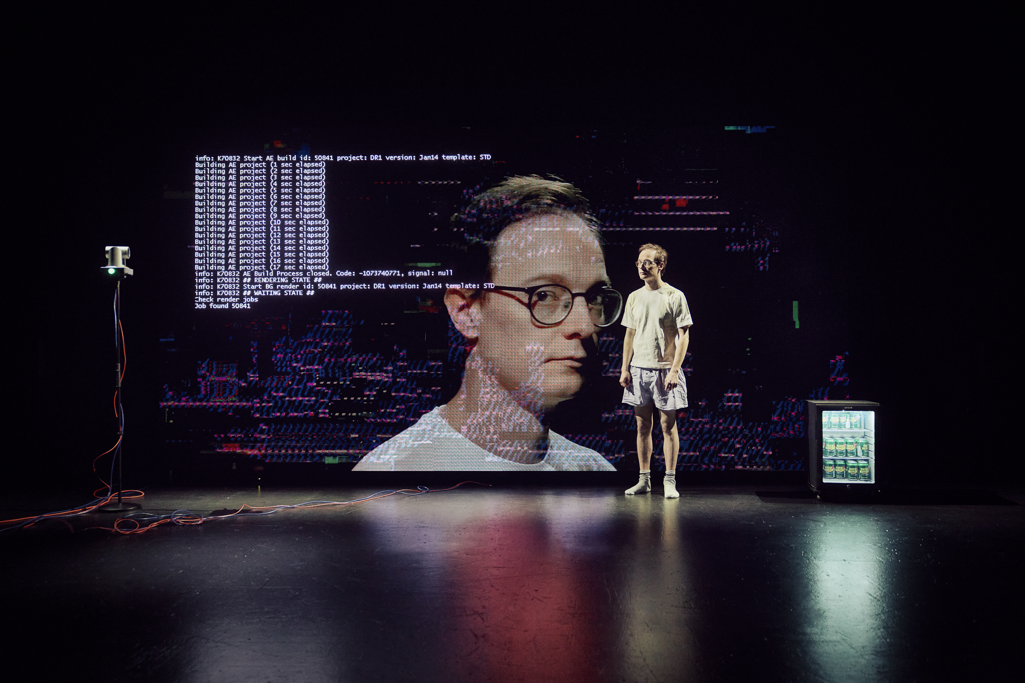 En mand i t-shirt og shorts står på en meget sort scene. En stor skærm bagved viser hans ansigt i nærbillede, samt kode fra computer. Fra forestillingen AI know you på Odense Teater