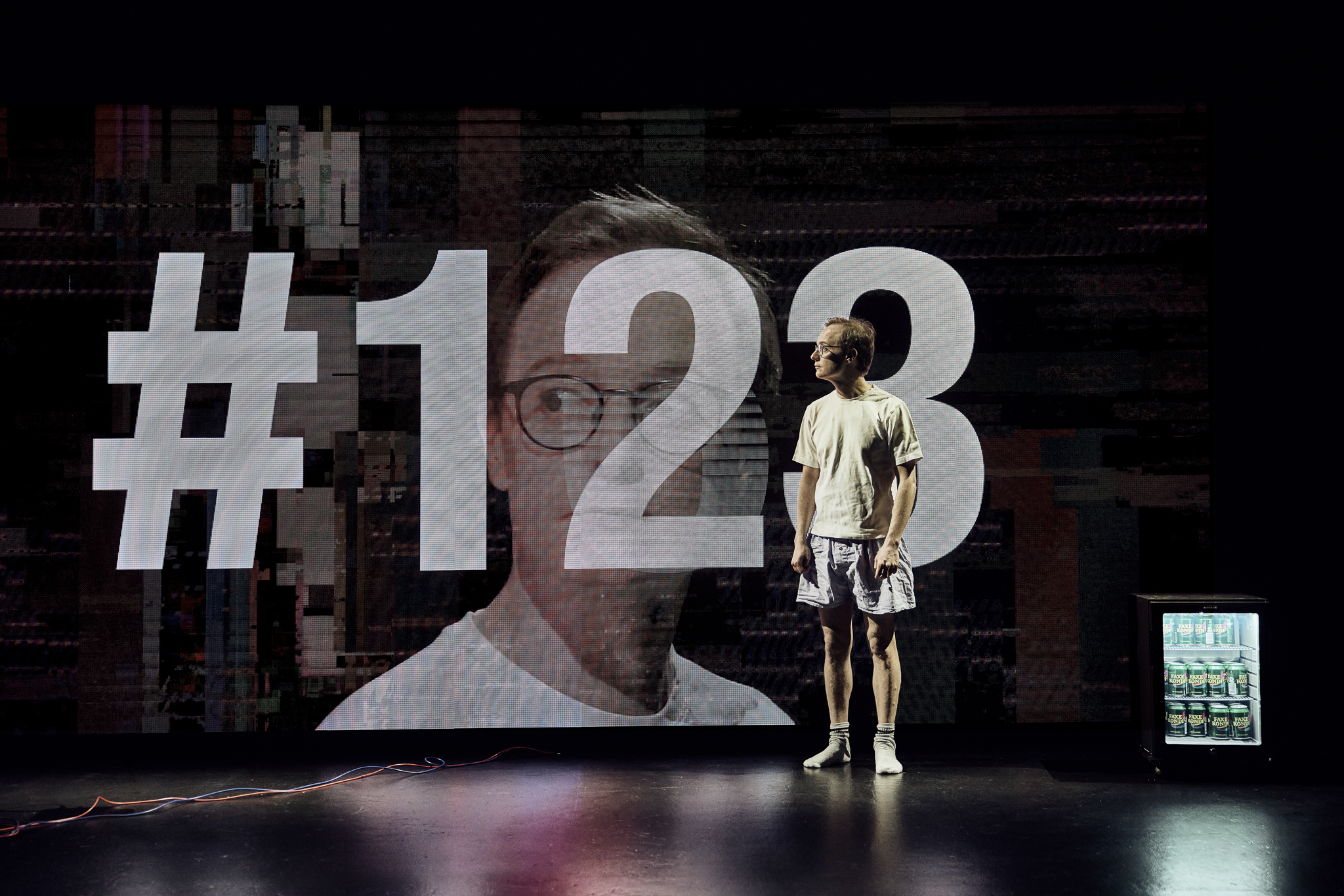 Mand i shorts og t-shirt står på en sort scene foran en skærm med hans ansigt i nærbillede og tallet 123. Fra forestillingen AI know you på Odense Teater