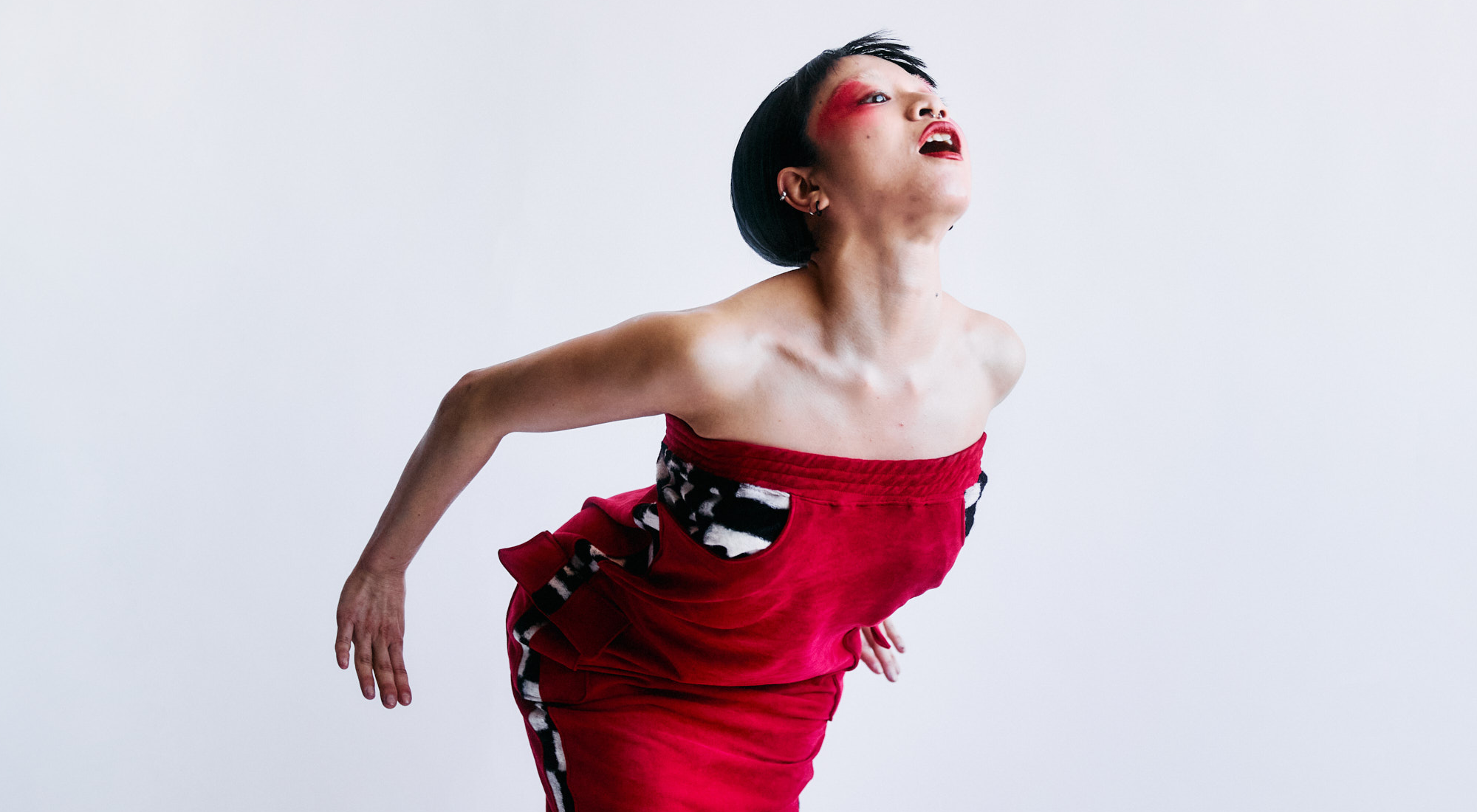 En asiatisk kvinde i rød kjole med bare skuldre rækker sig frem mod kameraet. Grå baggrund