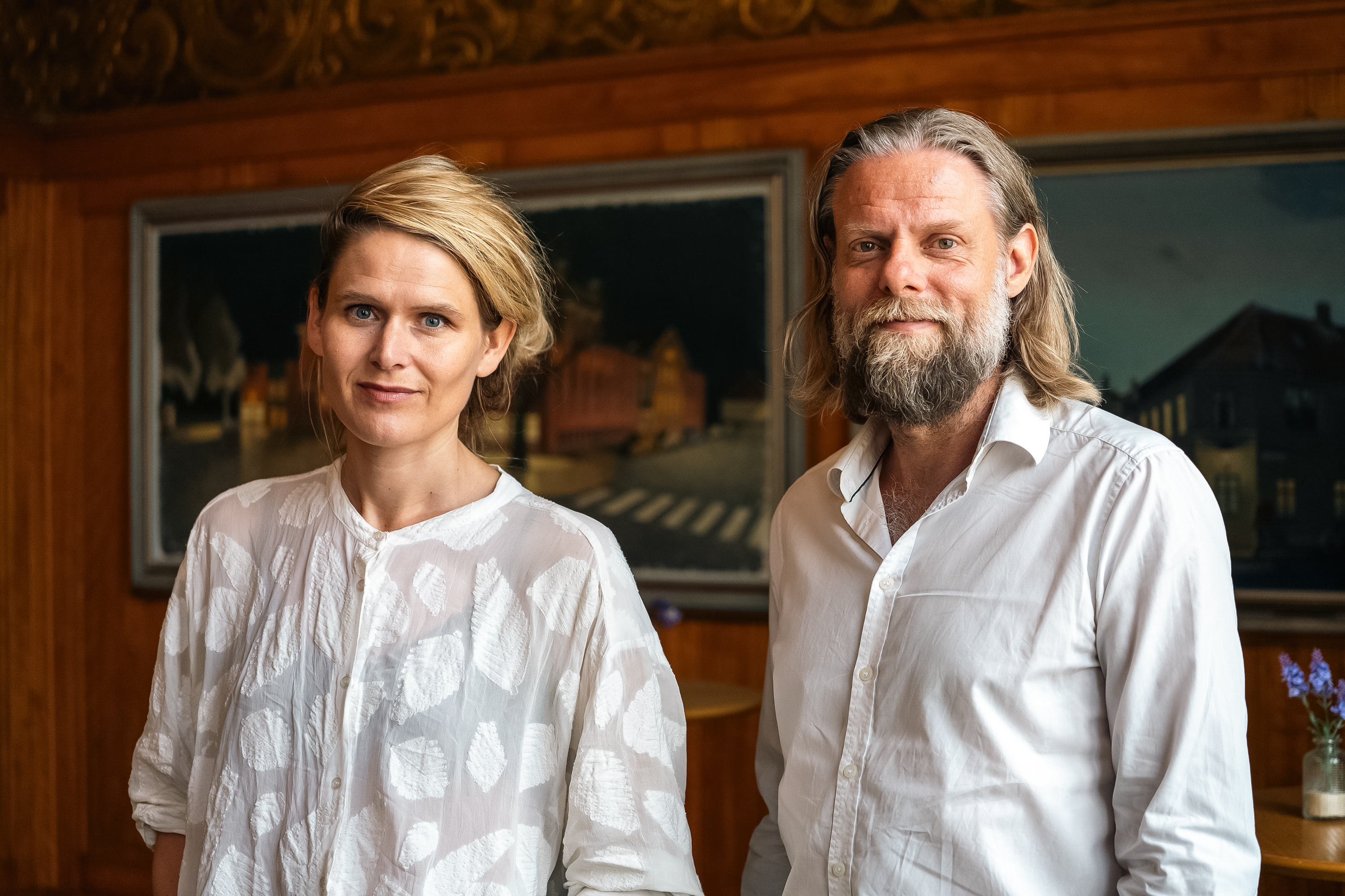 Liv Helm og Jacob Schjødt, begge i hvide skjorter står foran væg med gamle malerier på Odense Teater. 