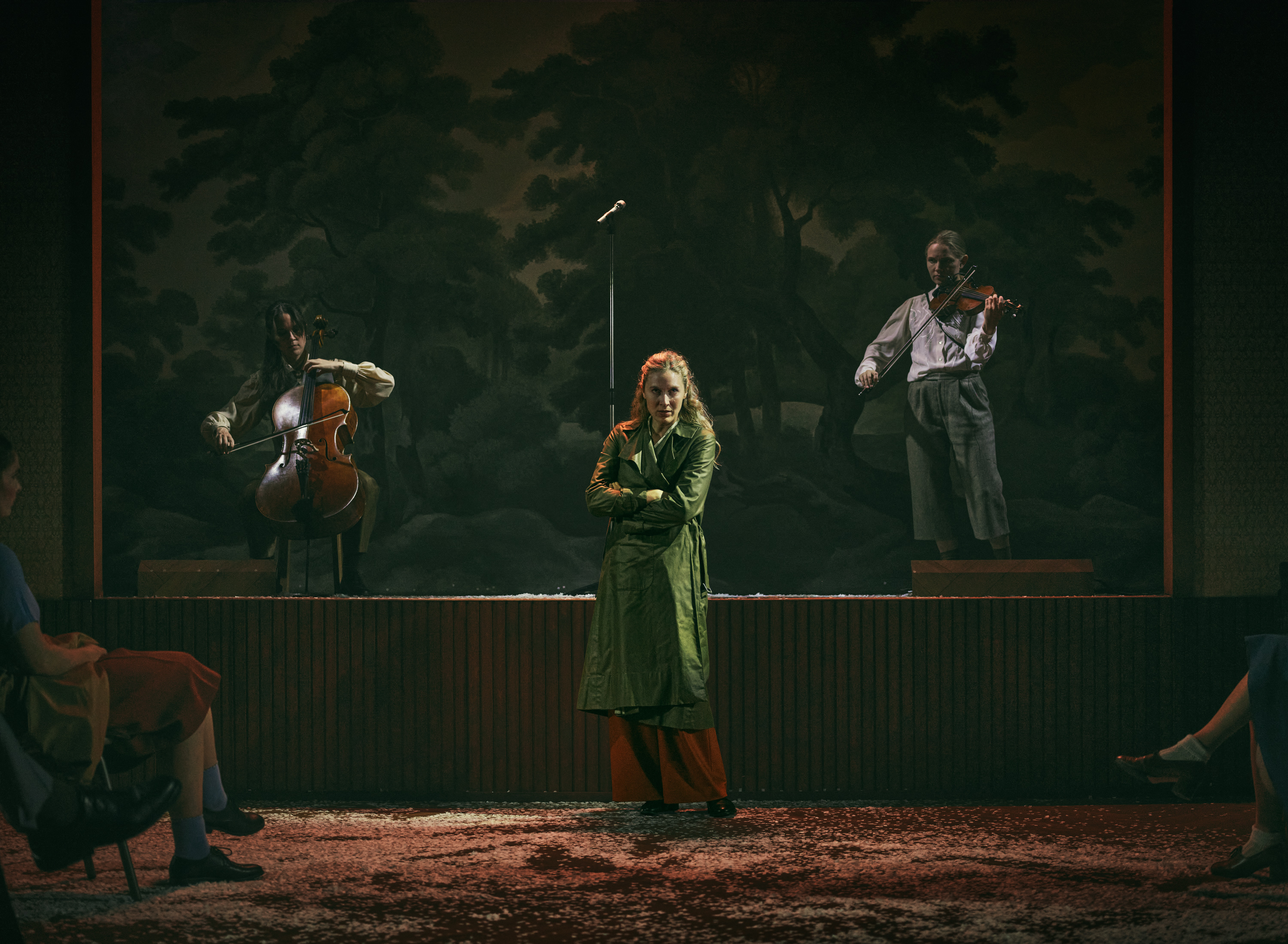 En kvinde står i front. I baggrund en scene, hvor der er en sidende kvinde med cello og en stående kvinde med violin. Billedet er mørkt. Fra forestillingen Alt i alt på Odense Teater