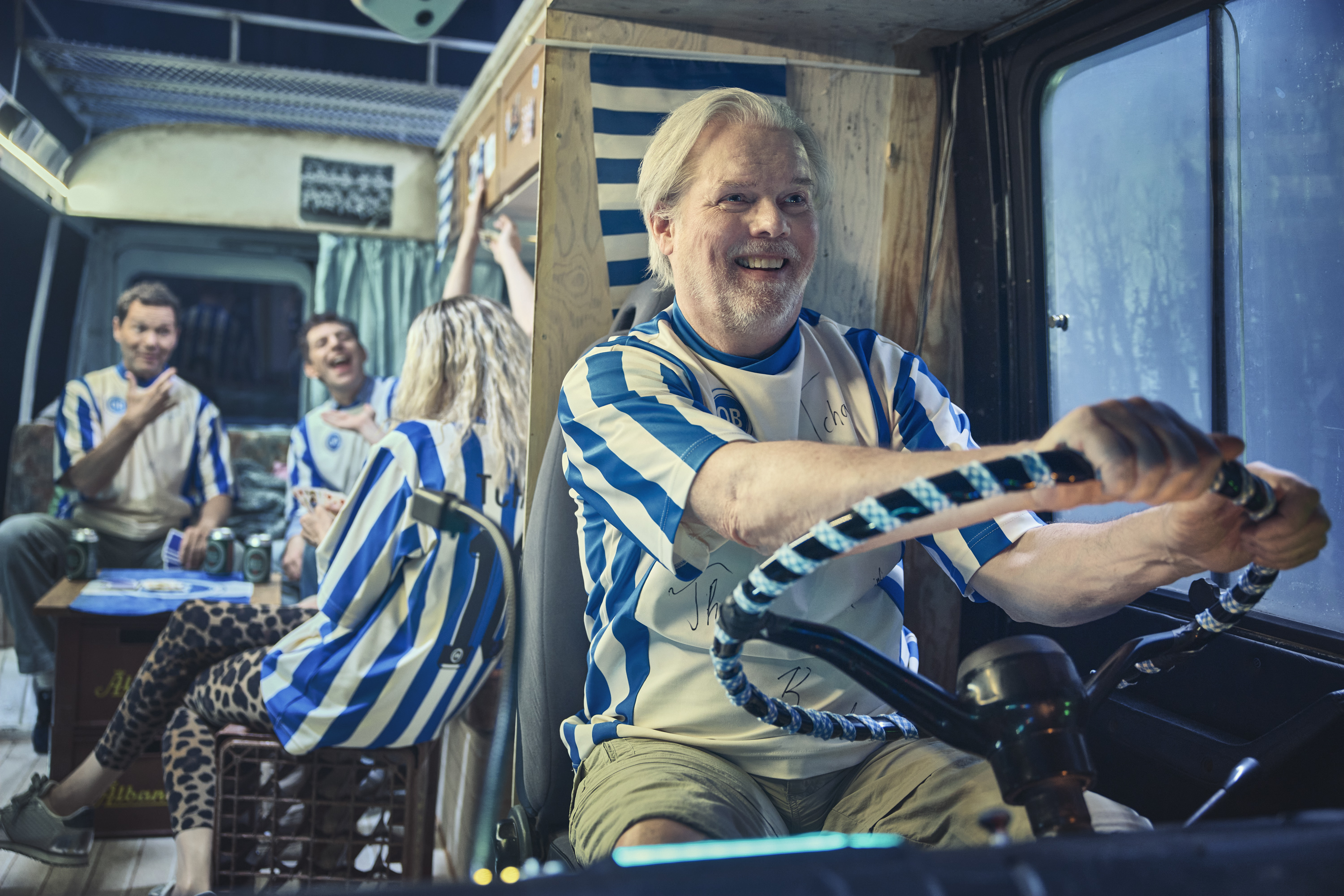 Mand i OB fodboldtrøje sidder bag rattet. I baggrund sidder tre mennesker og spiller kort. Fra teaterforestillingen Miraklet i Madrid på Odense Teater