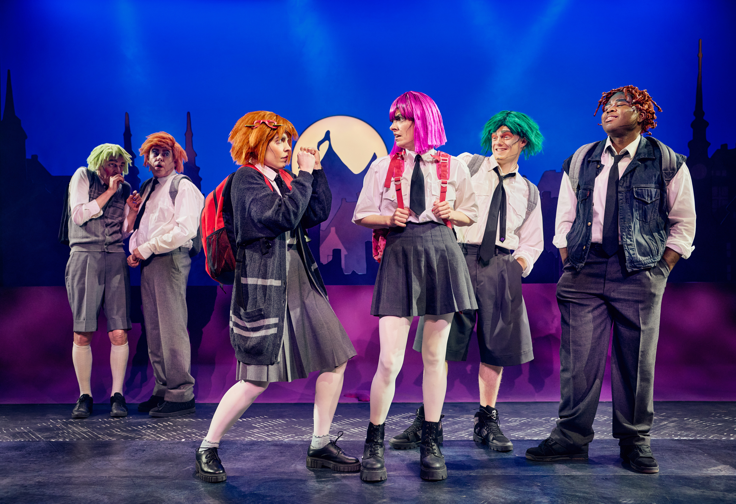 Pige med orange hår og skoleuniform går op i mod sine mobbere. 5 unge i skoleuniformer og farvet hår. Fra forestillingen Håndbog for superhelte på Odense Teater