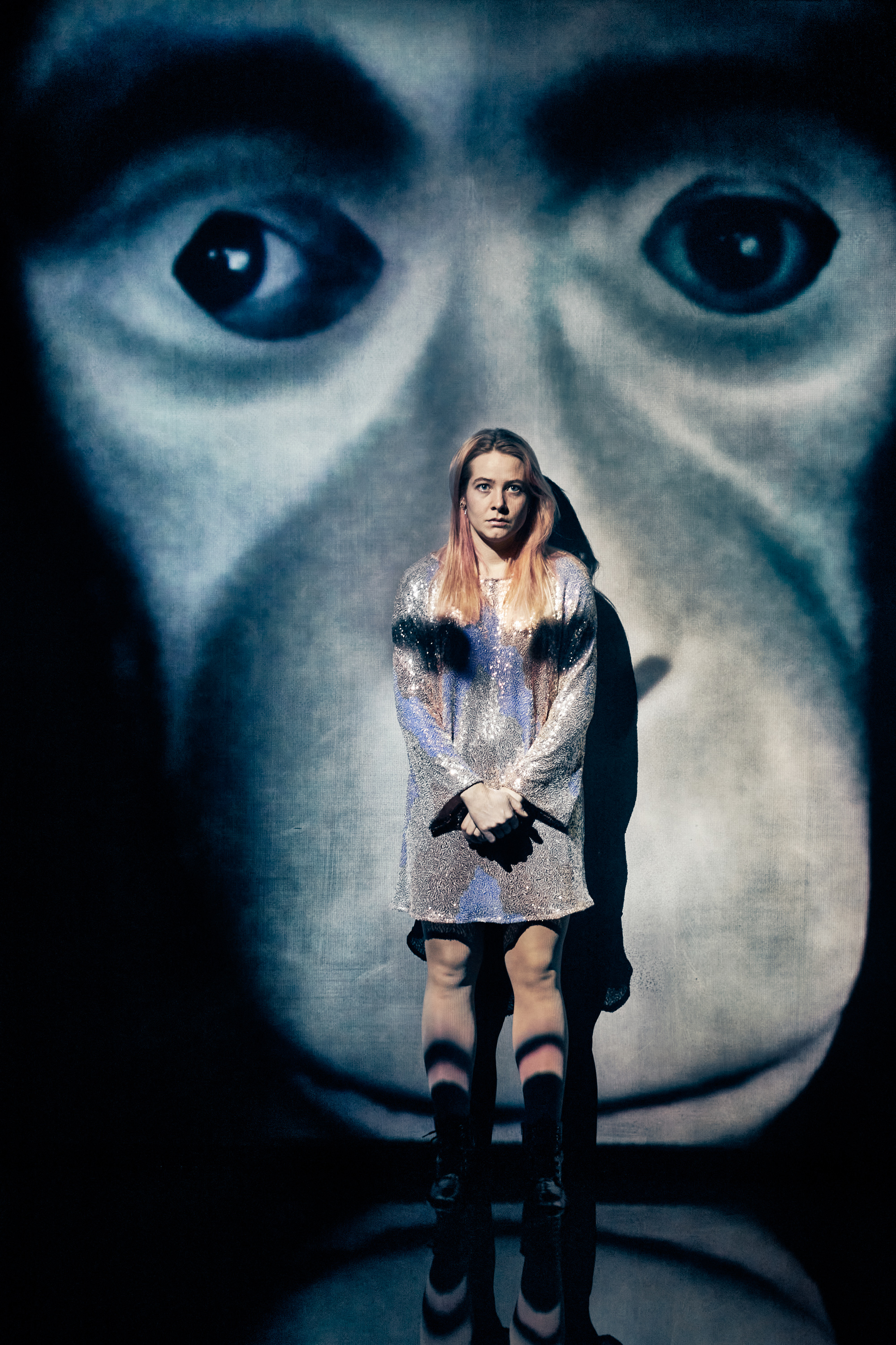 Ung kvinde i pailetkjole står op ad væg med billede af et abeansigt. Fra forestilling Maria på Odense Teater.