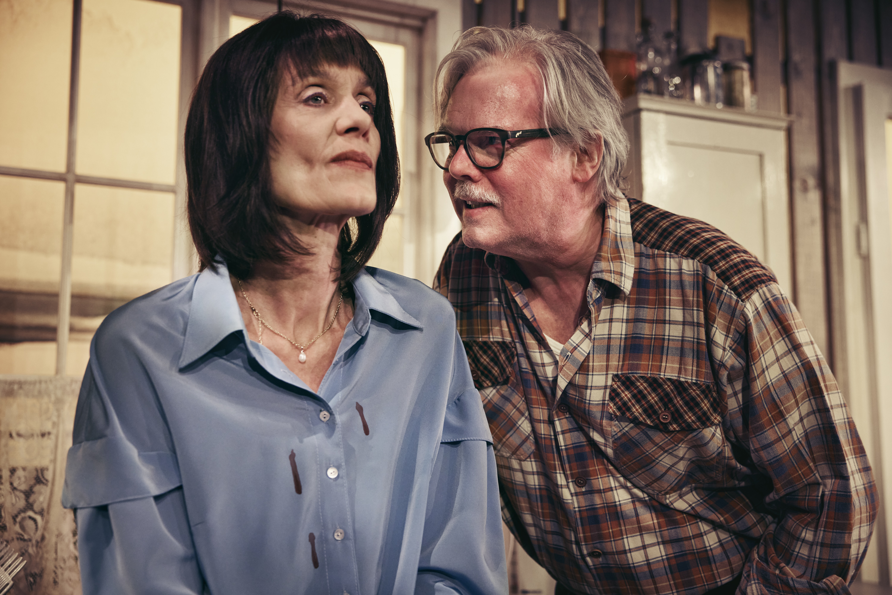 Ældre mand med mørke briller står tæt på mørkhåret kvinde i blå skjorte. Fra teaterforestilling Børnene på Odense Teater.