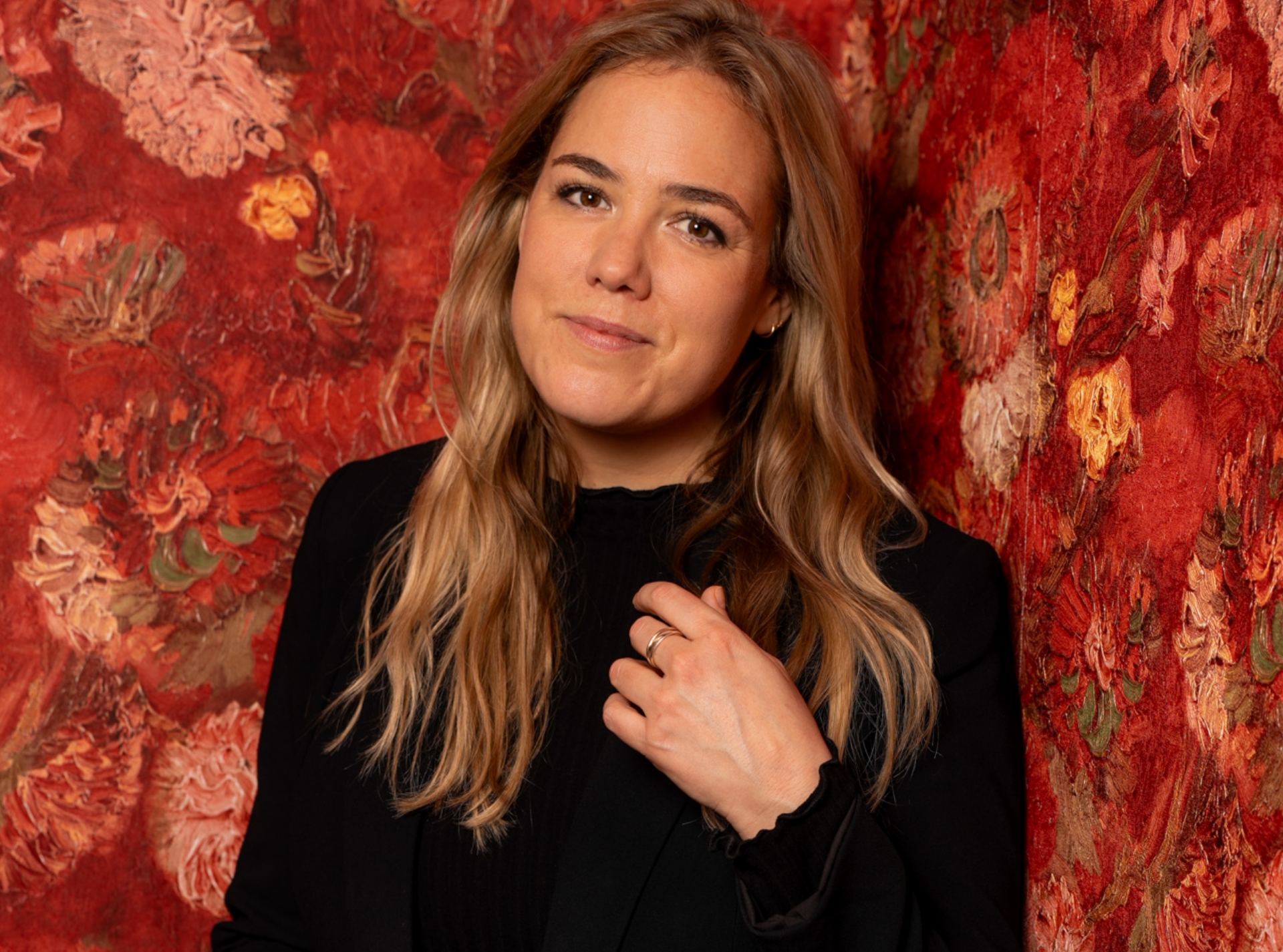 Portræt af Kvinde med langt hår iklædt sort bluse. Hun står op af væg med rødt mønstret tapet. Gry Jexen foredrag