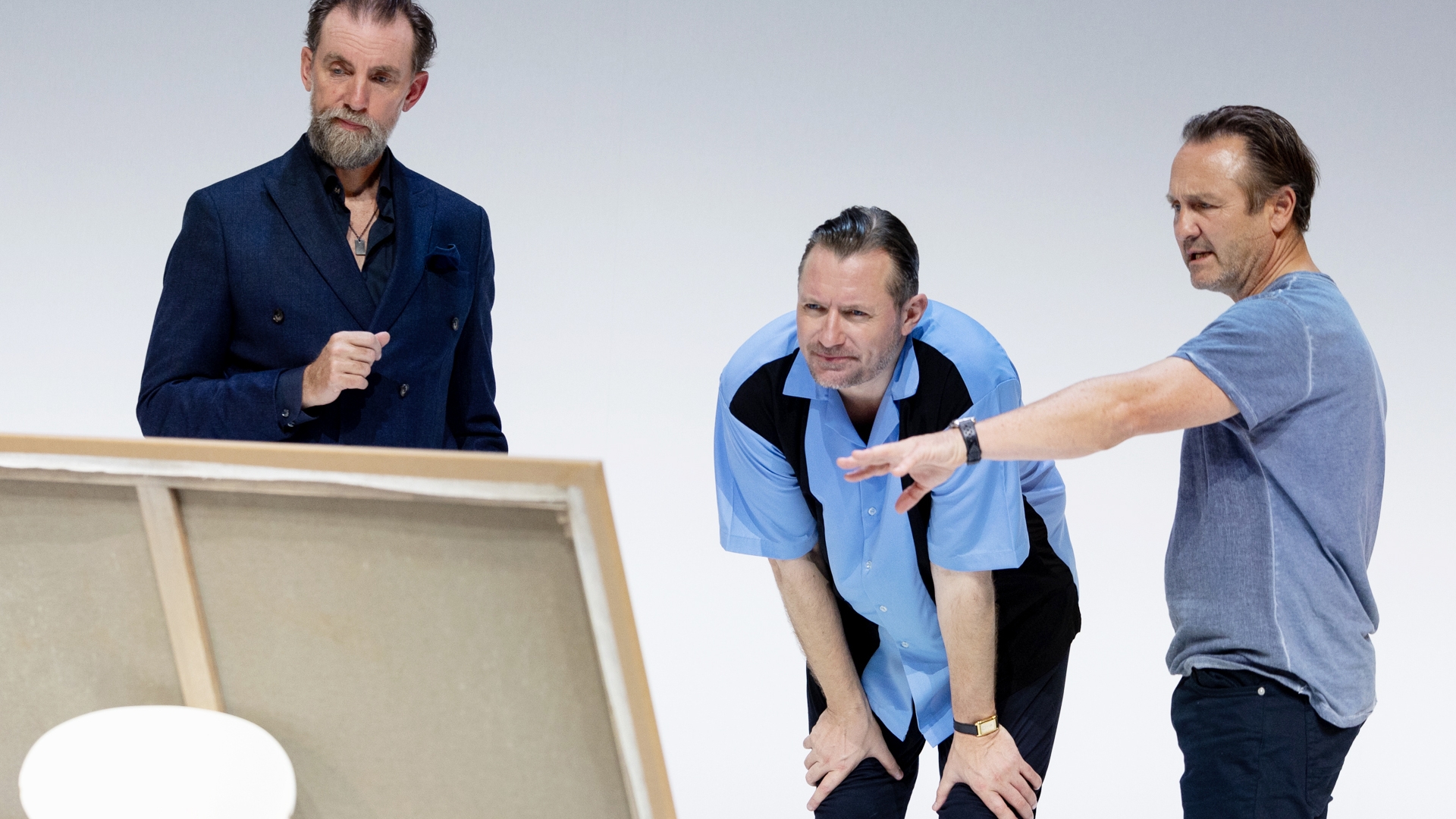 3 mænd ser vurderende på et maleri. Den ene mand peger på maleriet. Lys baggrund. Fra forestillingen Kunst