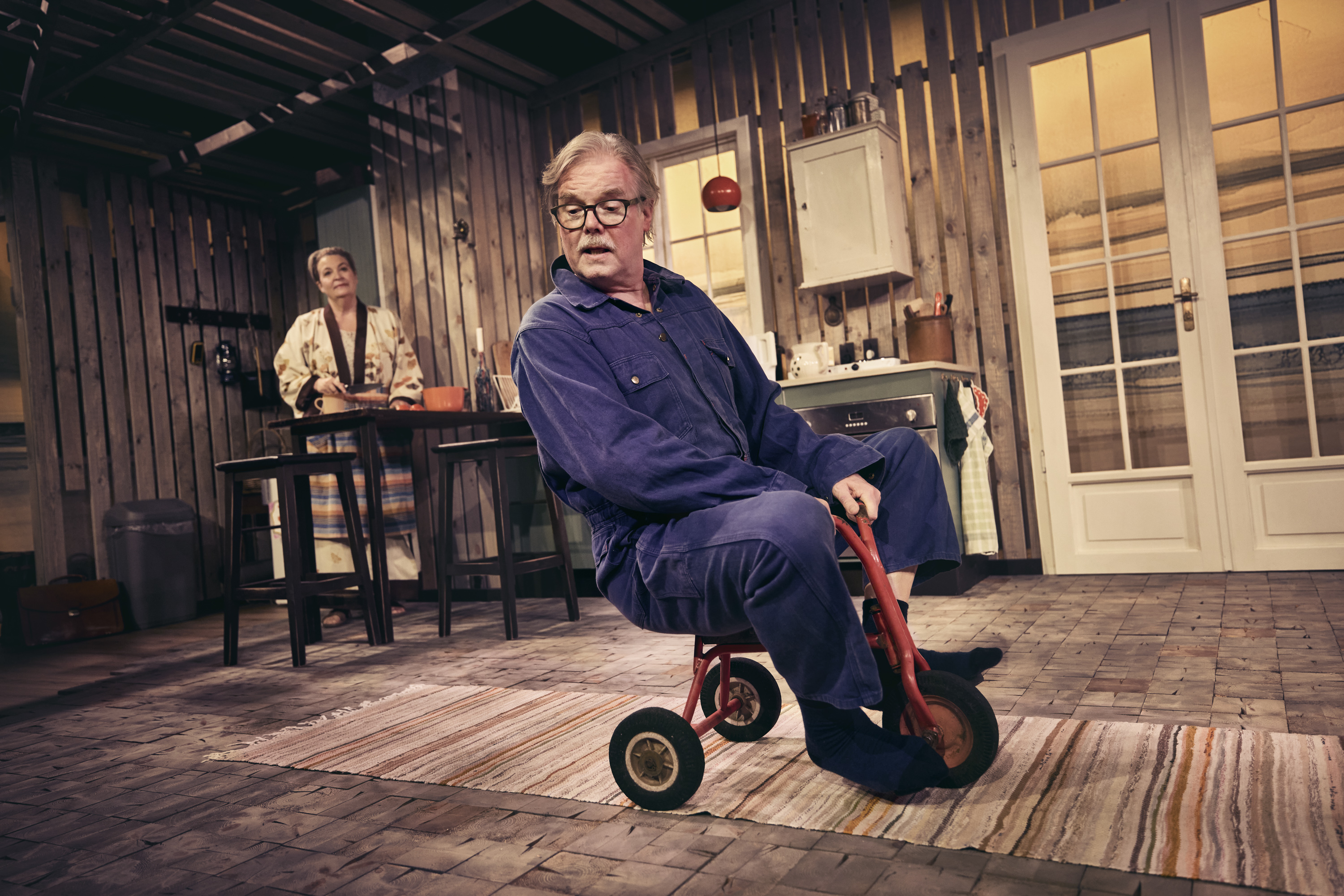 Mand i blå keddeldragt kører på lille rød trehjulet cykel i stue. Kvinde i baggrunden. Fra teaterforestilling Børnene på Odense Teater.