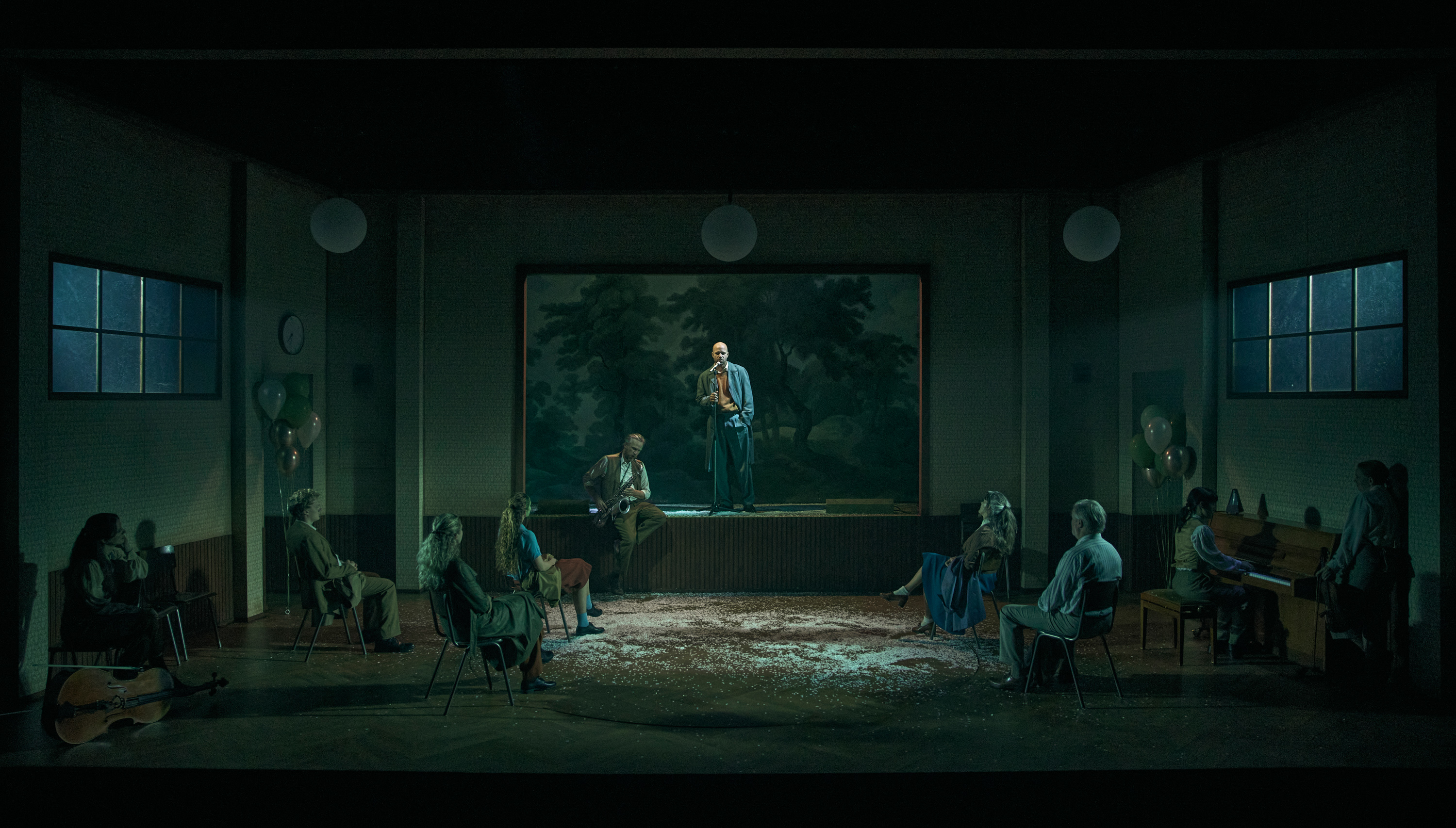 Flere mennesker i det der skal forestille et forsamlingshus. De sidder på stole og lytter til en mand der står på scenen. Fra forestillingen Alt i alt på Odense Teater
