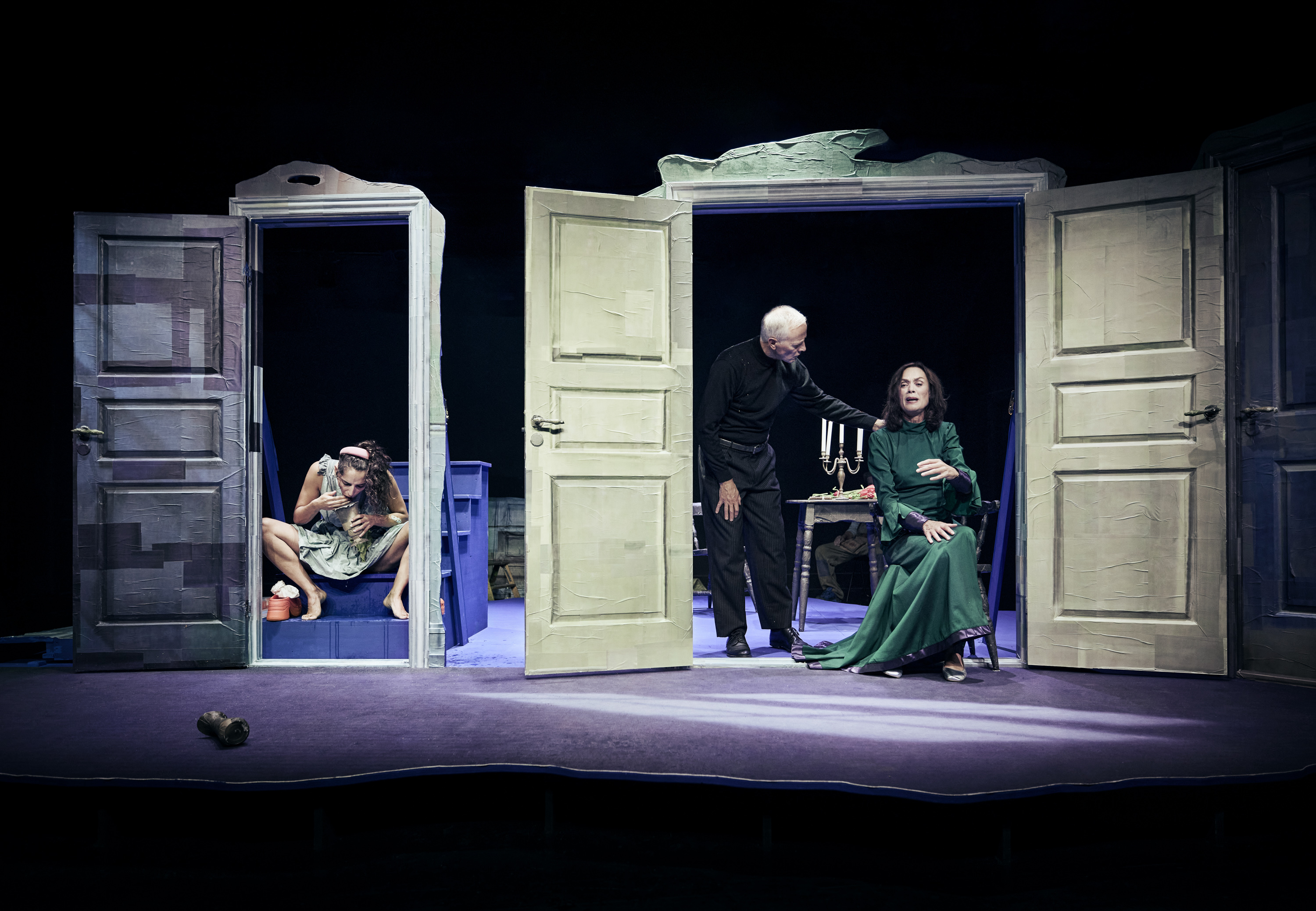 3 skuespillere på en scene. Scenografi er fra Gengangere på Odense Teater. Åbne døre. En kvinde sidder og spiser i den ene døråbning. I den anden sidder en kvinde i lang grøn kjole på stol, mens en mand står ved siden af med hånd på hendes skulder. 