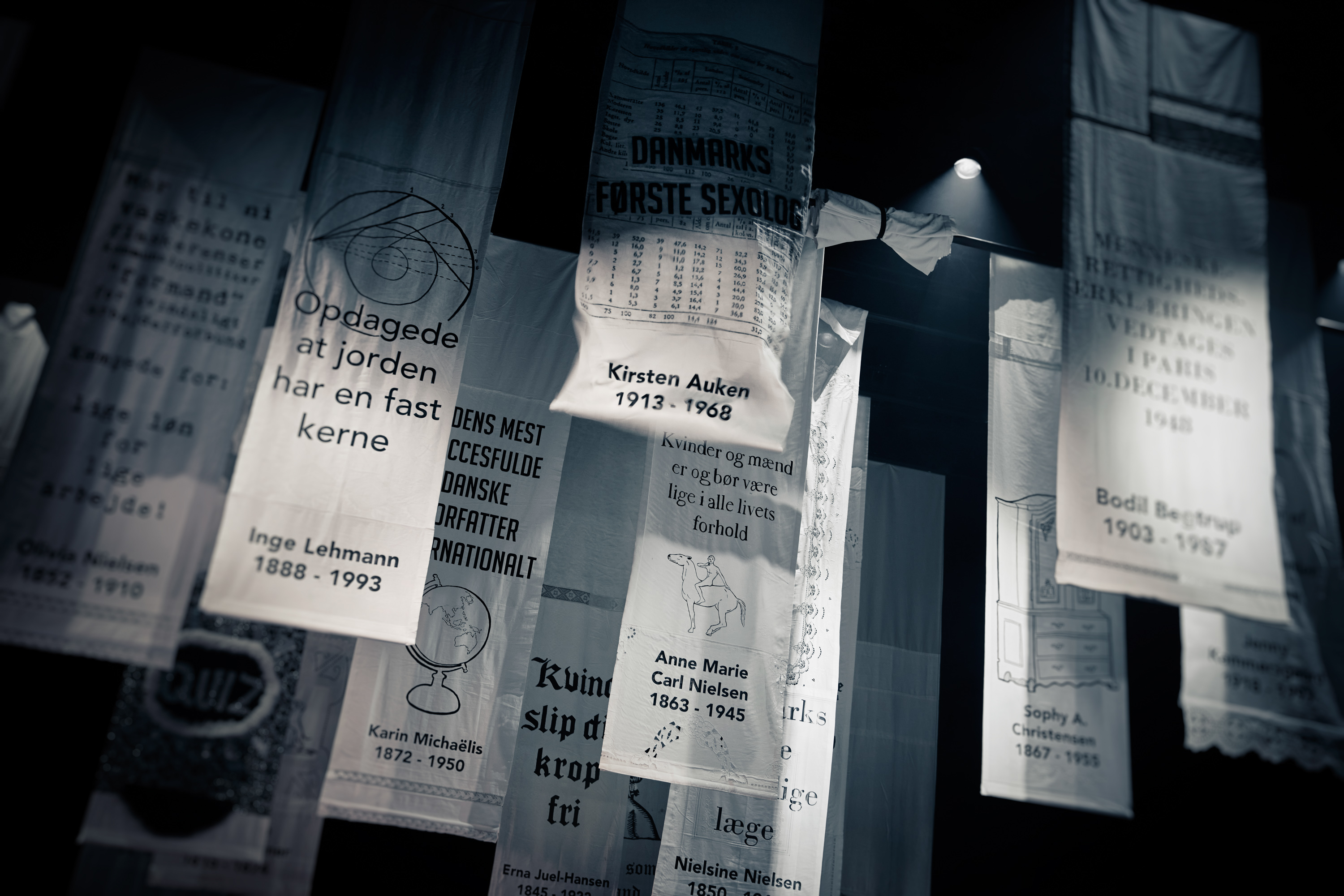 Hvide bannere hænger fra loftet. Hvert med tekst om forskellige historiske kvinder. Fra forestilling Kvinde kend din historie på Odense Teater