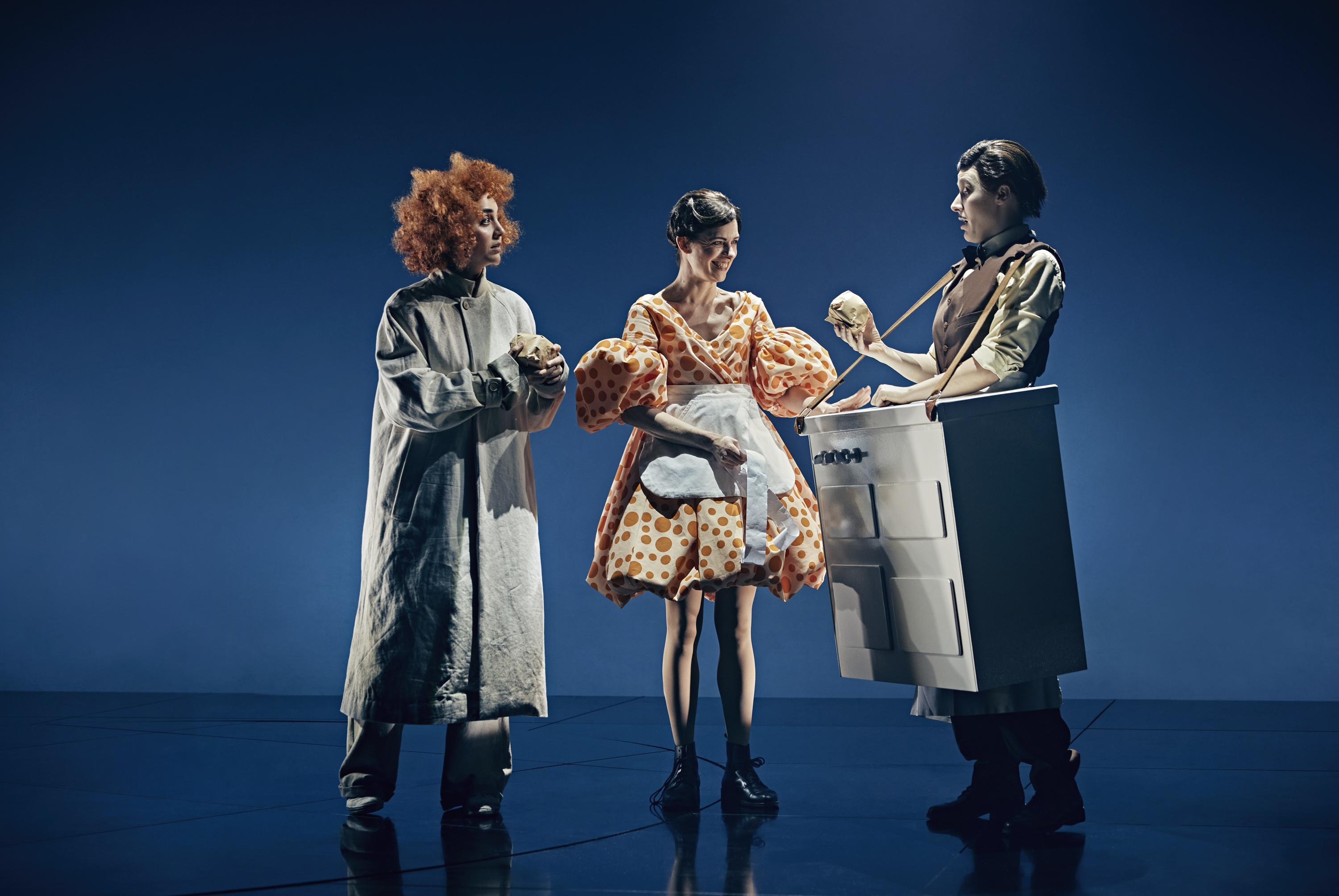 3 personer på billede med blå baggrund. En med rødt krøllet hår og lang frakke. En kvinde i kjole med orange prikker og pufærmer. De køber mad af mand der står med bod bundet til kroppen med strop om nakken.