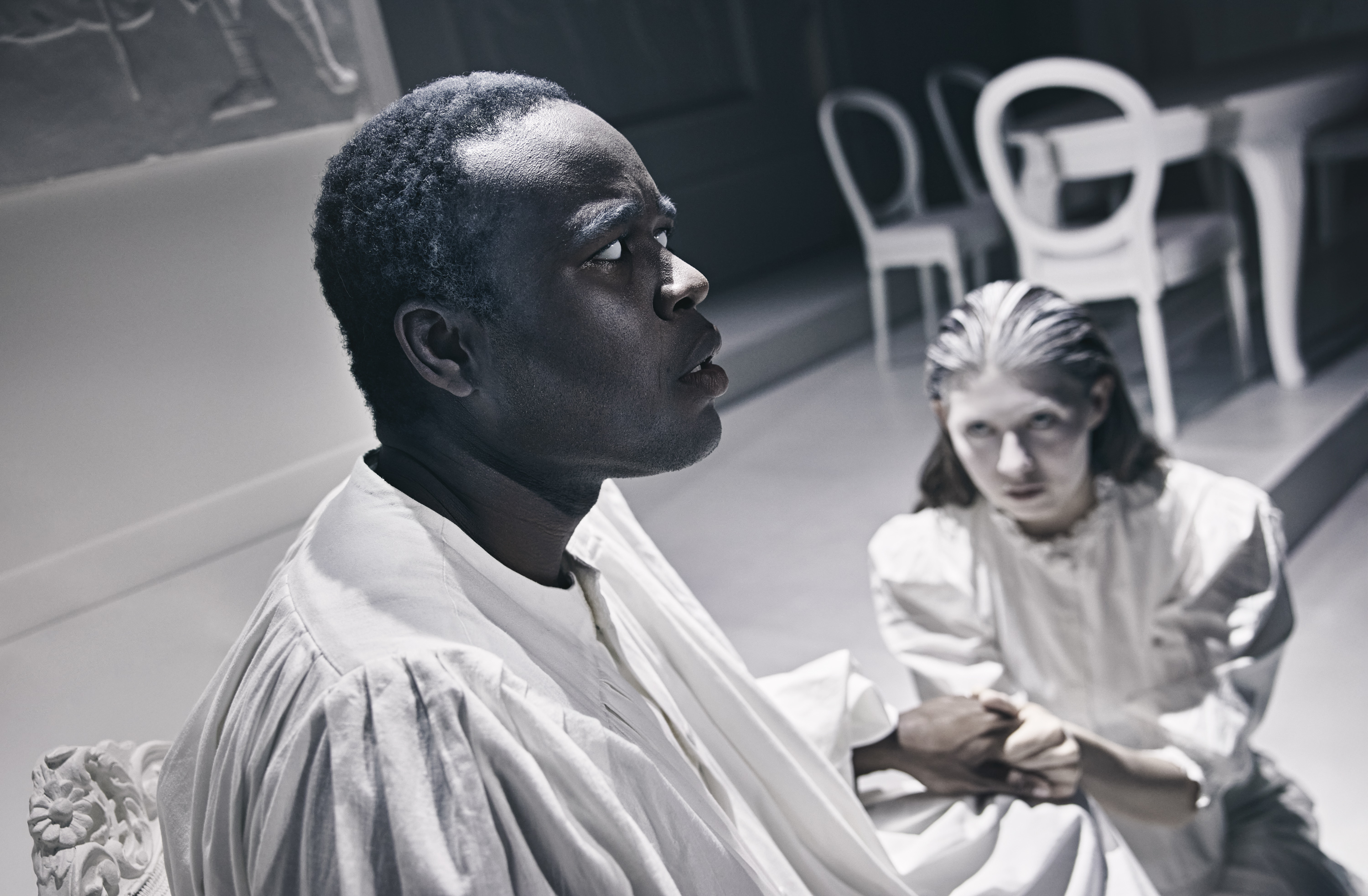 En sort mand klædt i hvidt sidder på stol. Ved hans side knæler en kvinde klædt i hvidt. Fra forestilling Resten kender du fra film på Odense Teater