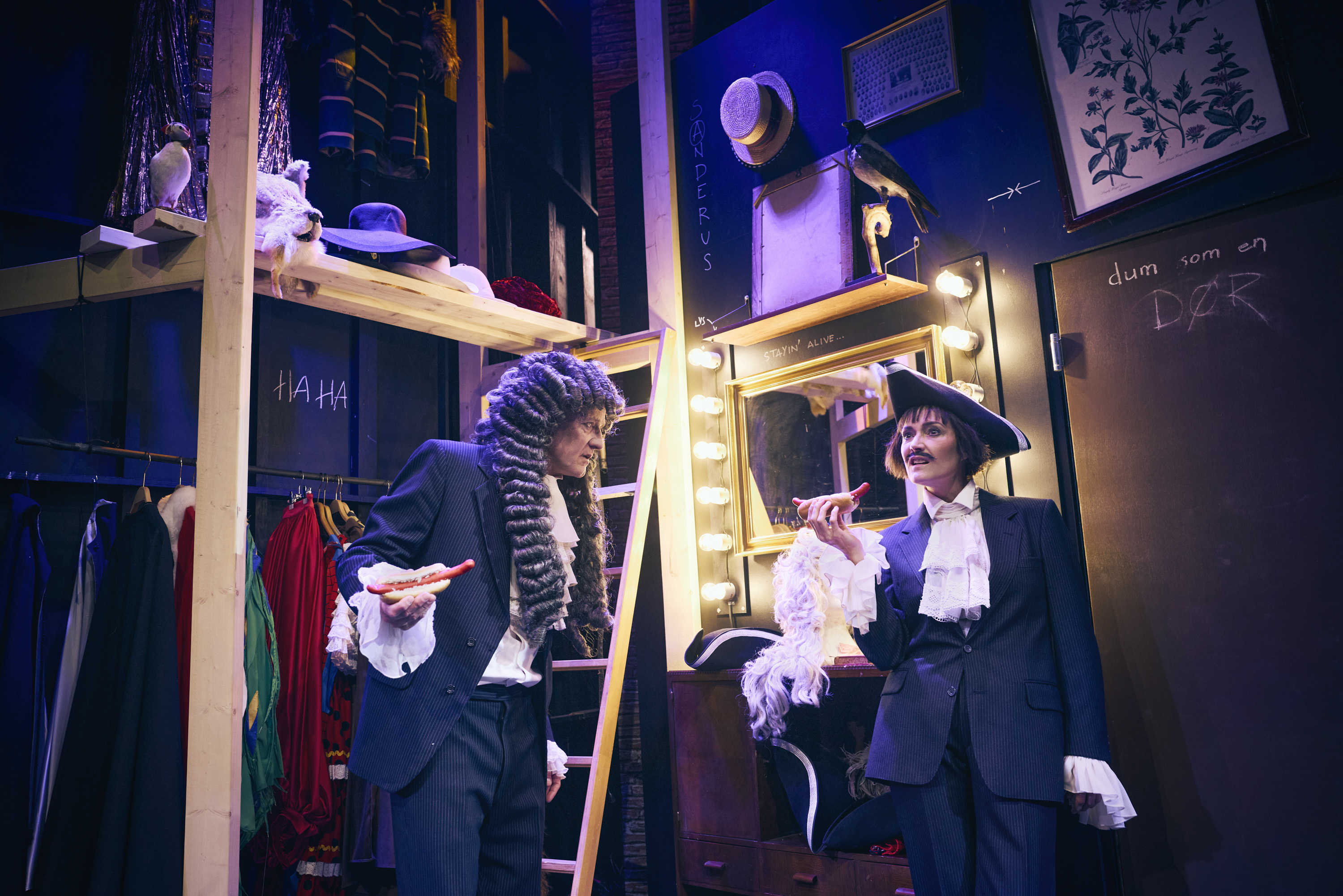 To mennesker i kostumer med lang paryk og kalvkrøs. De spiser hotdogs. Fra teaterforestillingen Den politiske kandestøber på Odense Teater