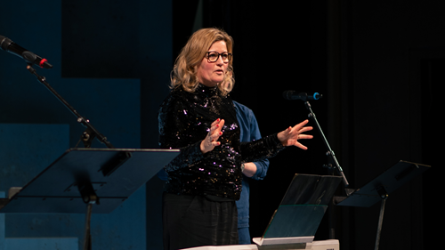 Kvinde med langt lyst hår står på scene foran mikrofon til Syng sammen på Odense Teater