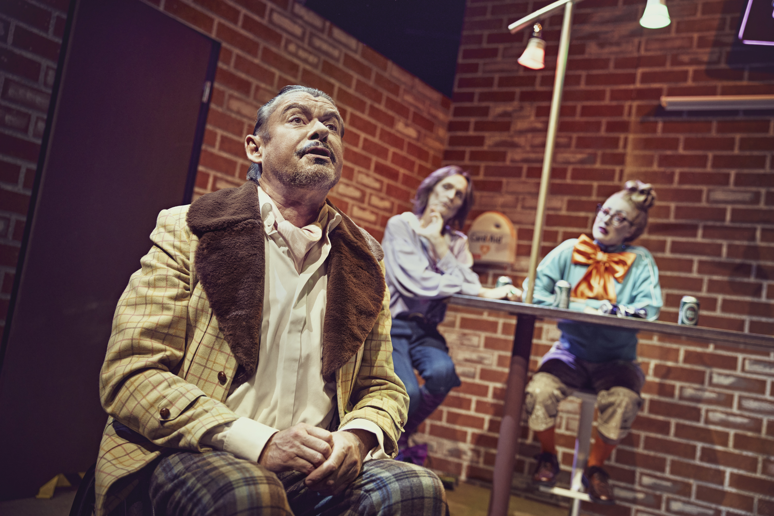 En mand sidder på en stol med et opgivende udtryk. To personer sidder ved en bar i baggrunden. Fra teaterforestillingen Den politiske kandestøber på Odense Teater