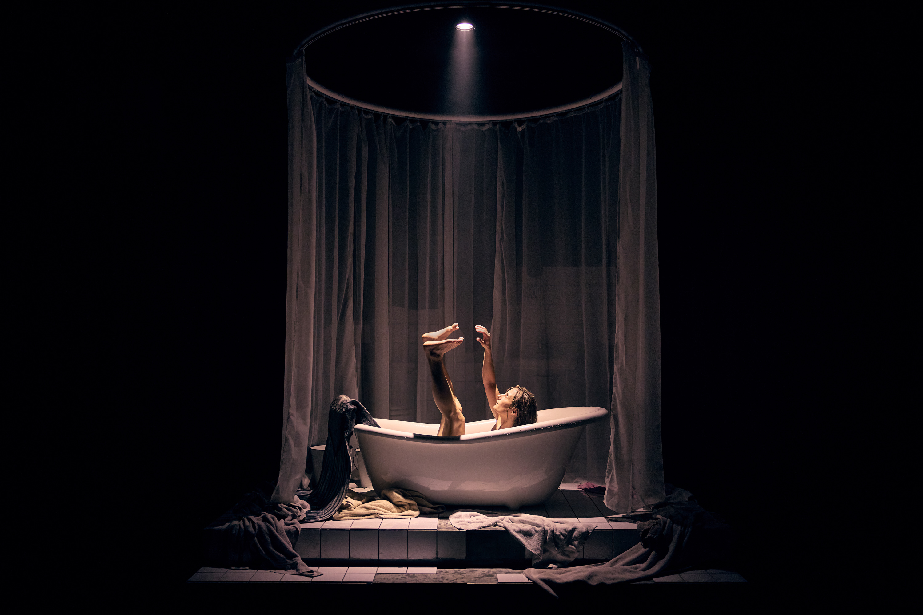 En mørk scene hvor der står et badekar på gulvet. Spotlys ned fra loftet. I badekaret ligger en nøgen kvinde med benene strakt op i luften. Fra forestillingen Fruen fra havet på Odesense Teater. 