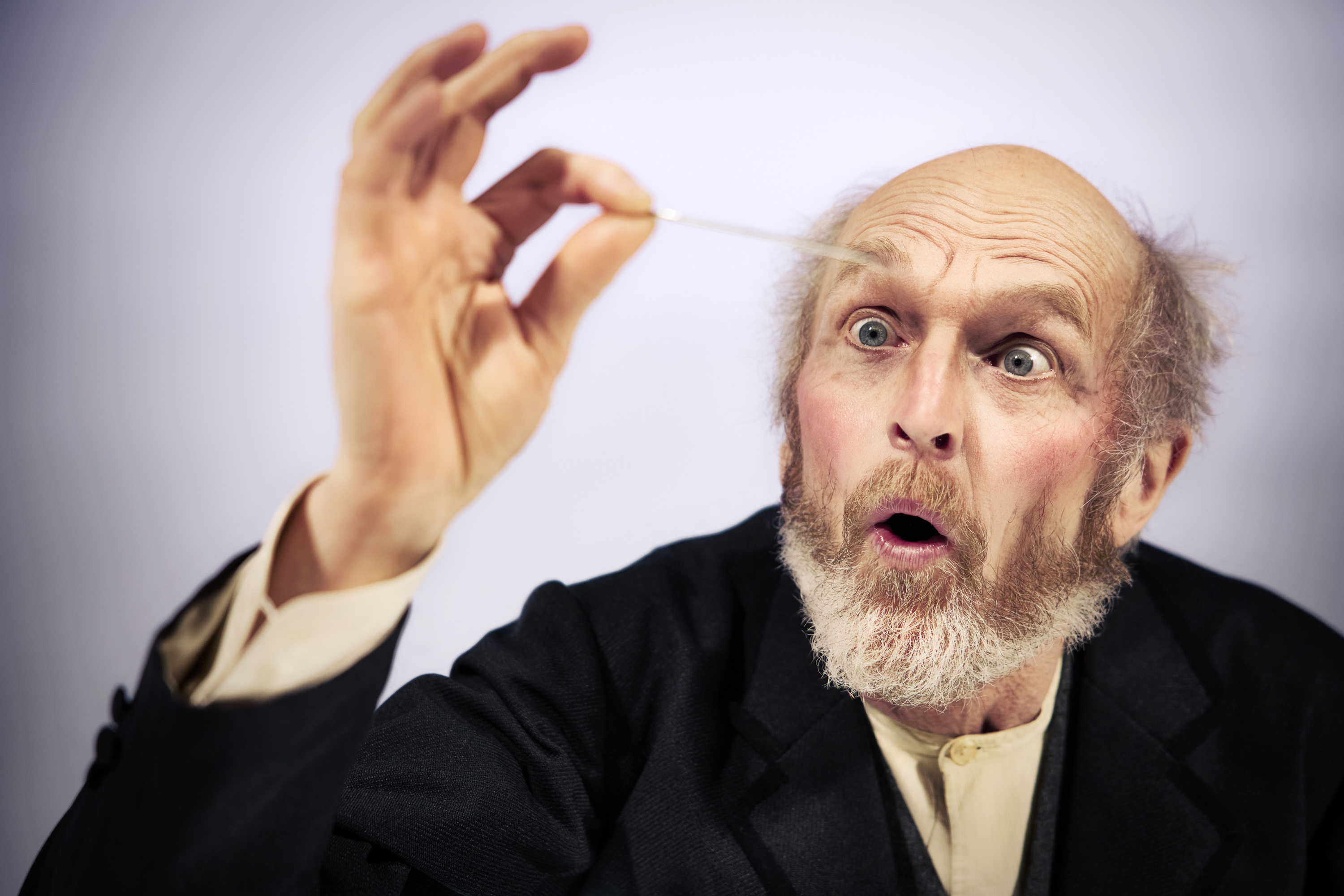 Ældre mand med skæg ser overrasket på fjer han holder mellem to fingre. Fra Teaterforestilling Klodshans på Odense Teater