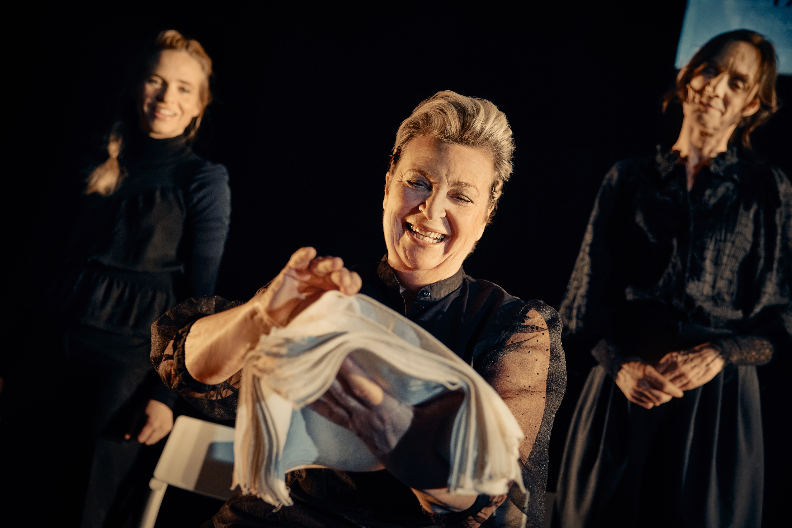 En glad kvinde i front. Hun holder hvidt stof. 2 kvinder i baggrunden klædt i sort. Fra forestilling Kvinde kend din historie på Odense Teater