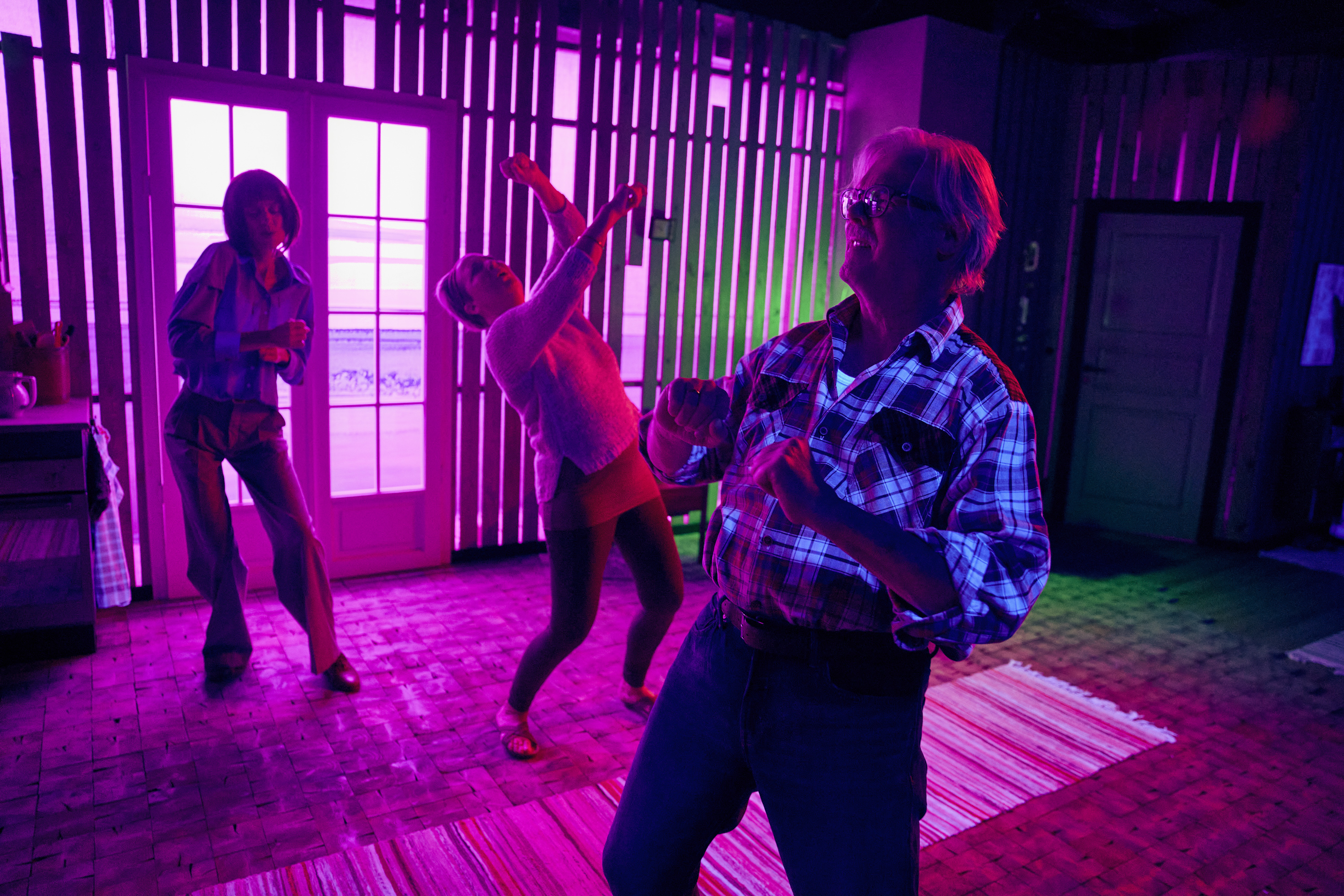 To kvinder og en mand danser i stue med et lilla lysskær. Fra teaterforestilling Børnene på Odense Teater.