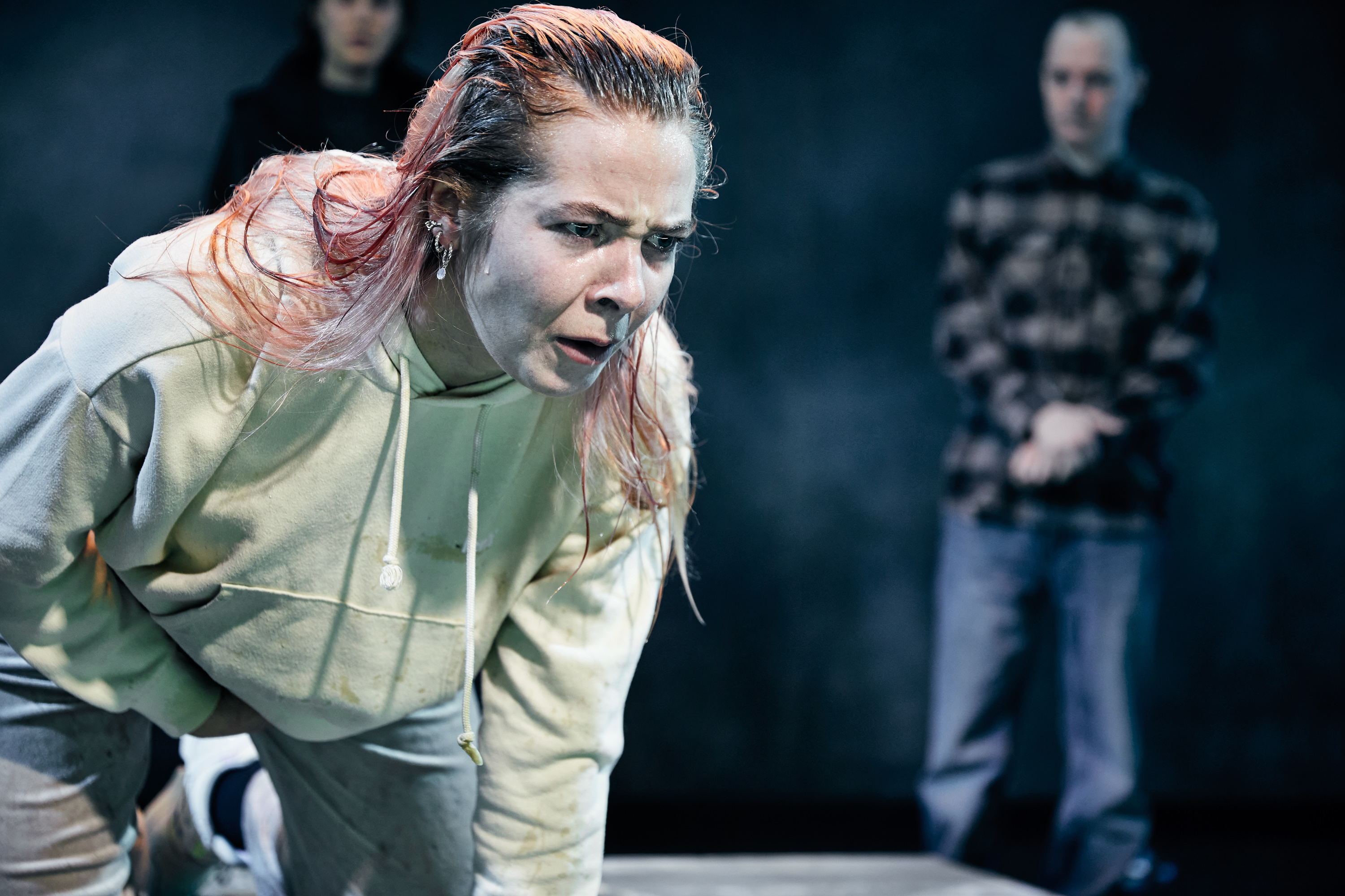 Ung kvinde på knæ med vådt hår og i tydelige smerter. 2 mænd i baggrunden. Fra forestilling Maria på Odense Teater