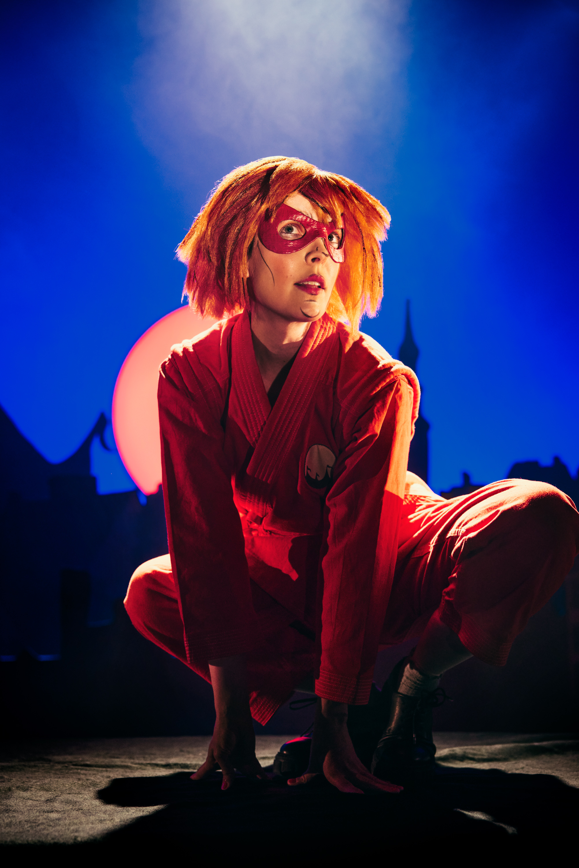 En pige med orange hår, rødt tøj og en rød superheltemaske sidder på hug. Fra forestillingen Håndbog for superhelte på Odense Teater