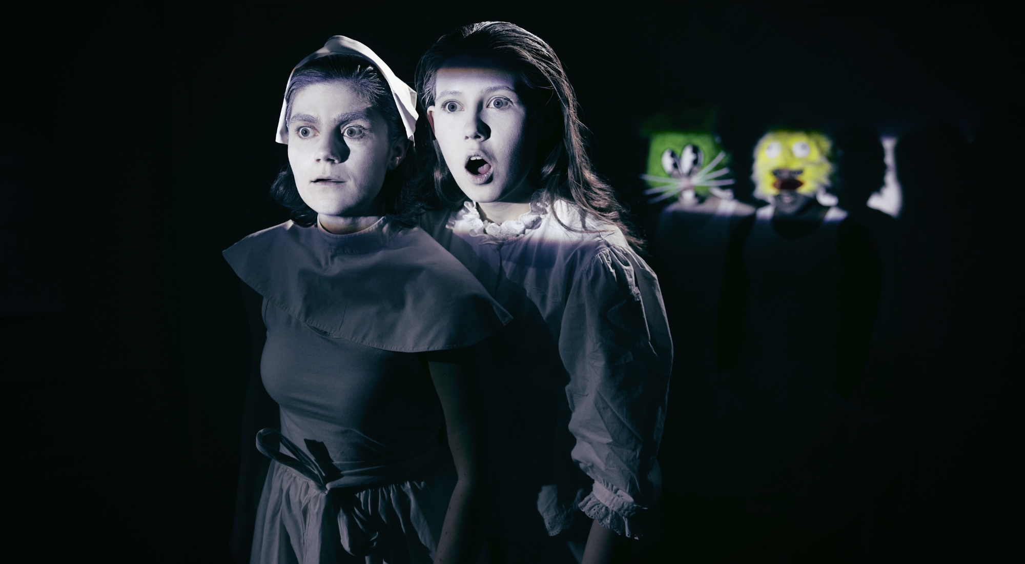 To piger i sort tøj, malet hvide i ansigterne står i helt sort rum. De har måbende ansigtsudtryk. I baggrunden ses ansigter af to tegneseriefigurer i grøn og gul. Fra Odense Teater